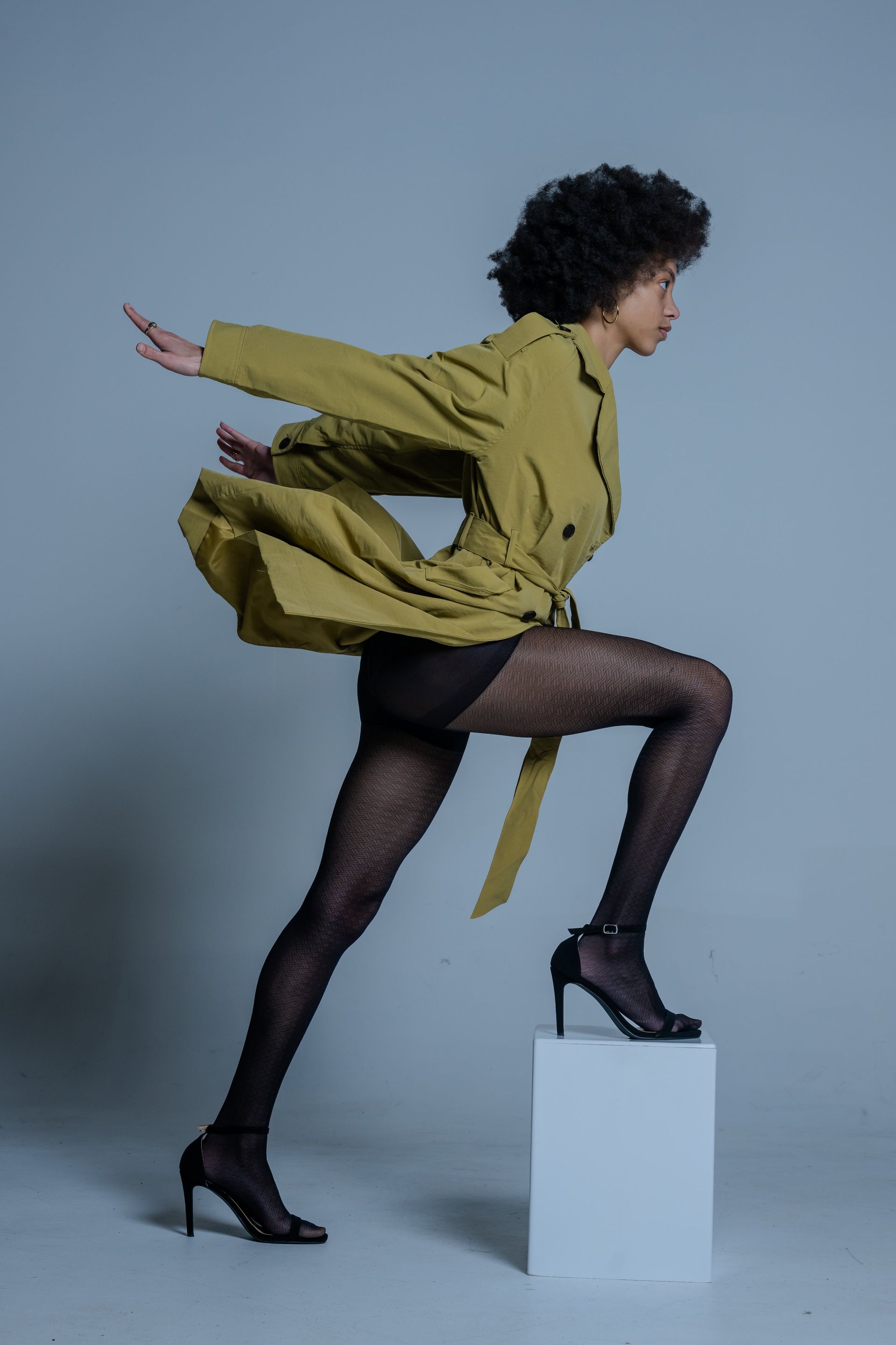 Person in olive jacket and black tights poses mid-step on a white block against a gray background.