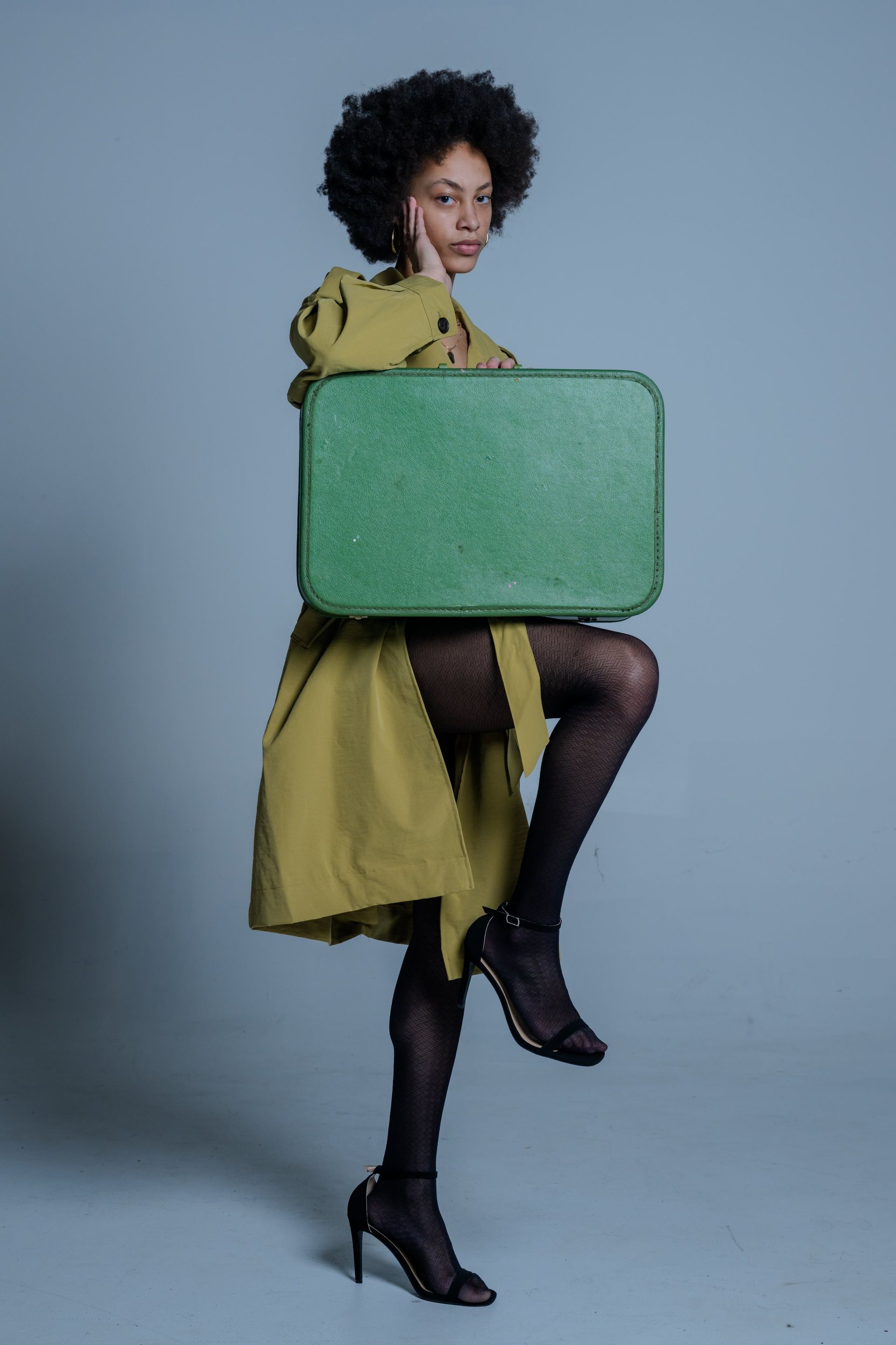 Person in a yellow dress and black tights holds a large green suitcase against a gray background.