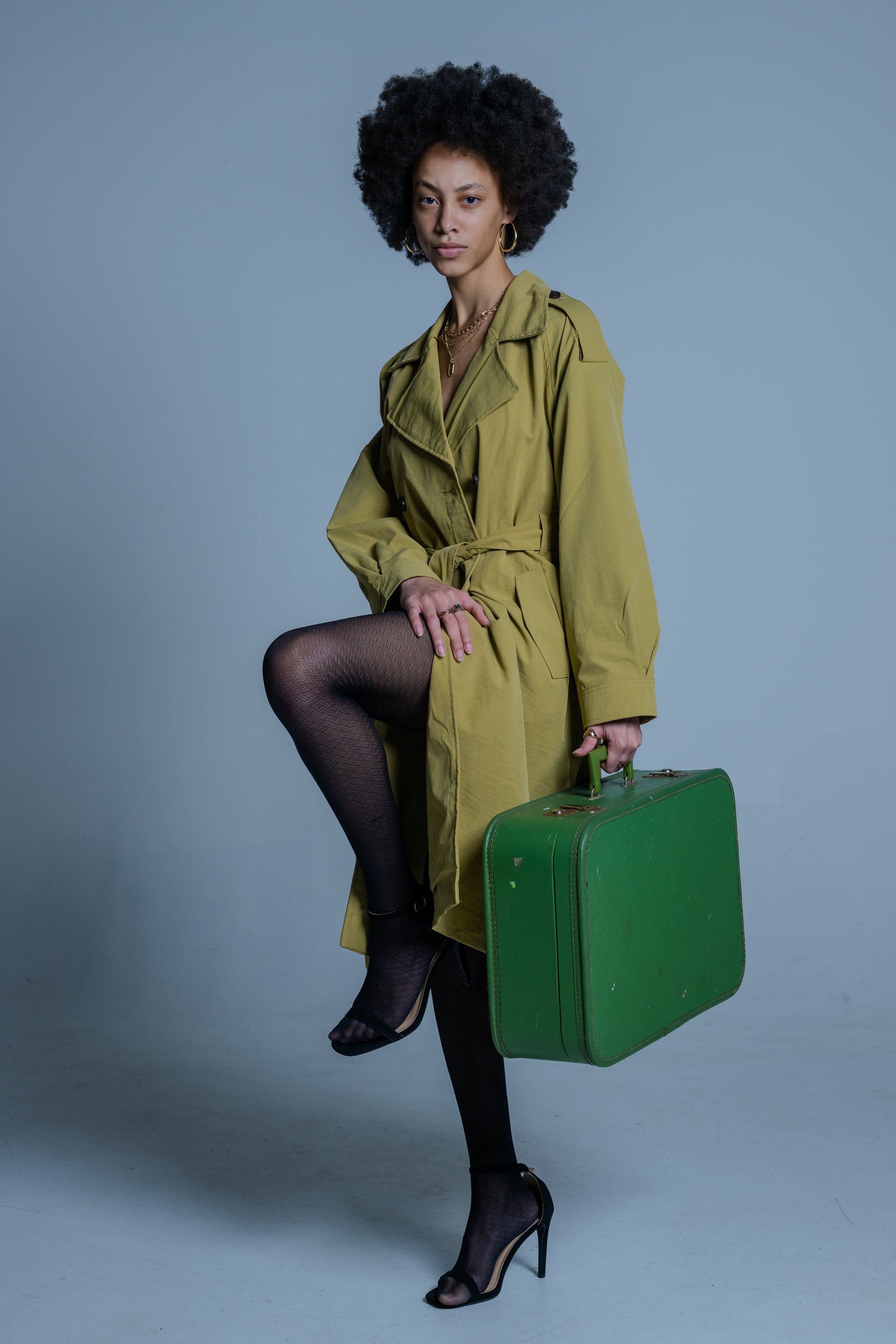 Person in an olive coat and black heels poses with a green suitcase against a gray backdrop.