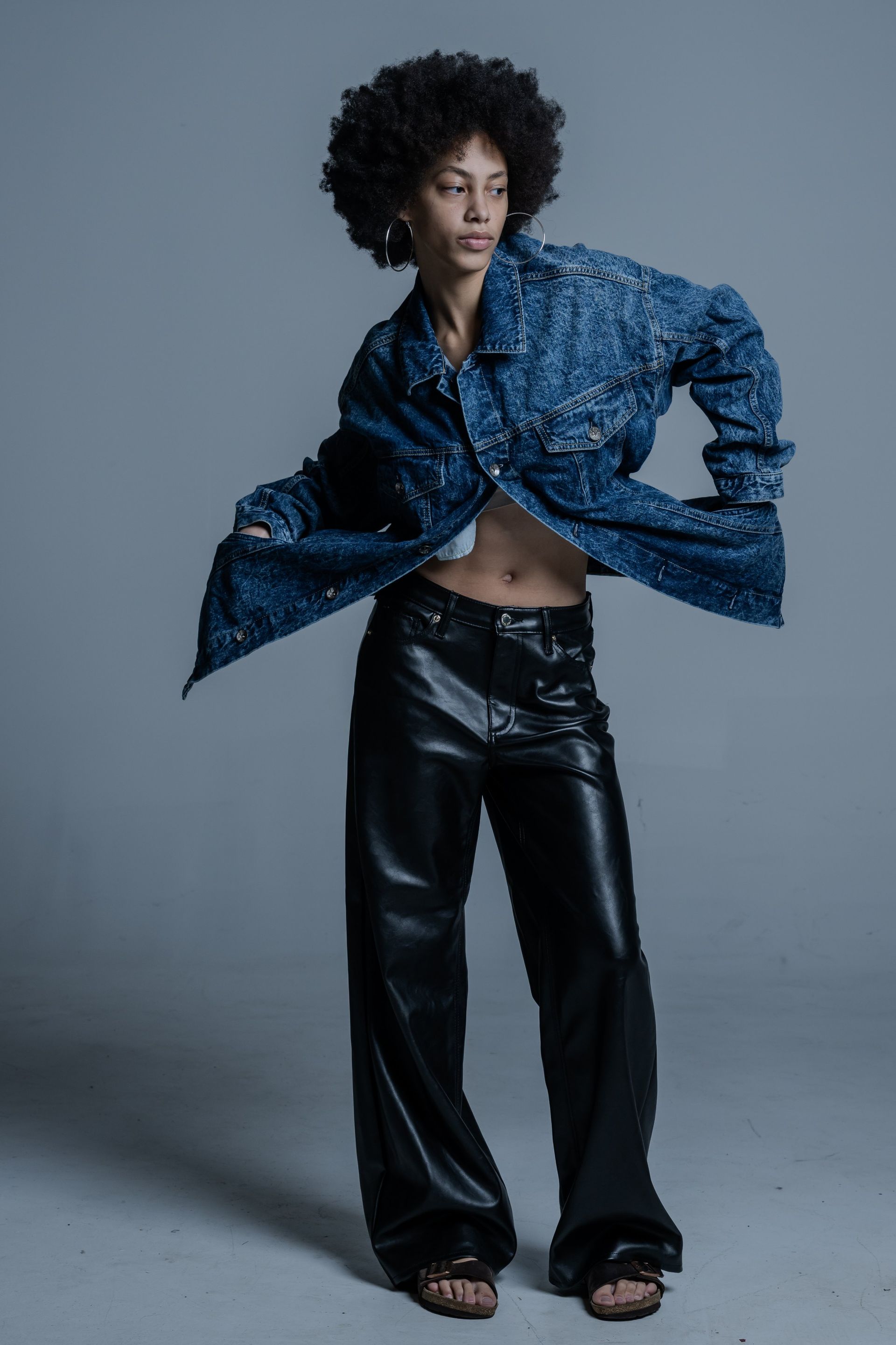 Person in a blue patterned shirt and black pants posing against a gray backdrop.
