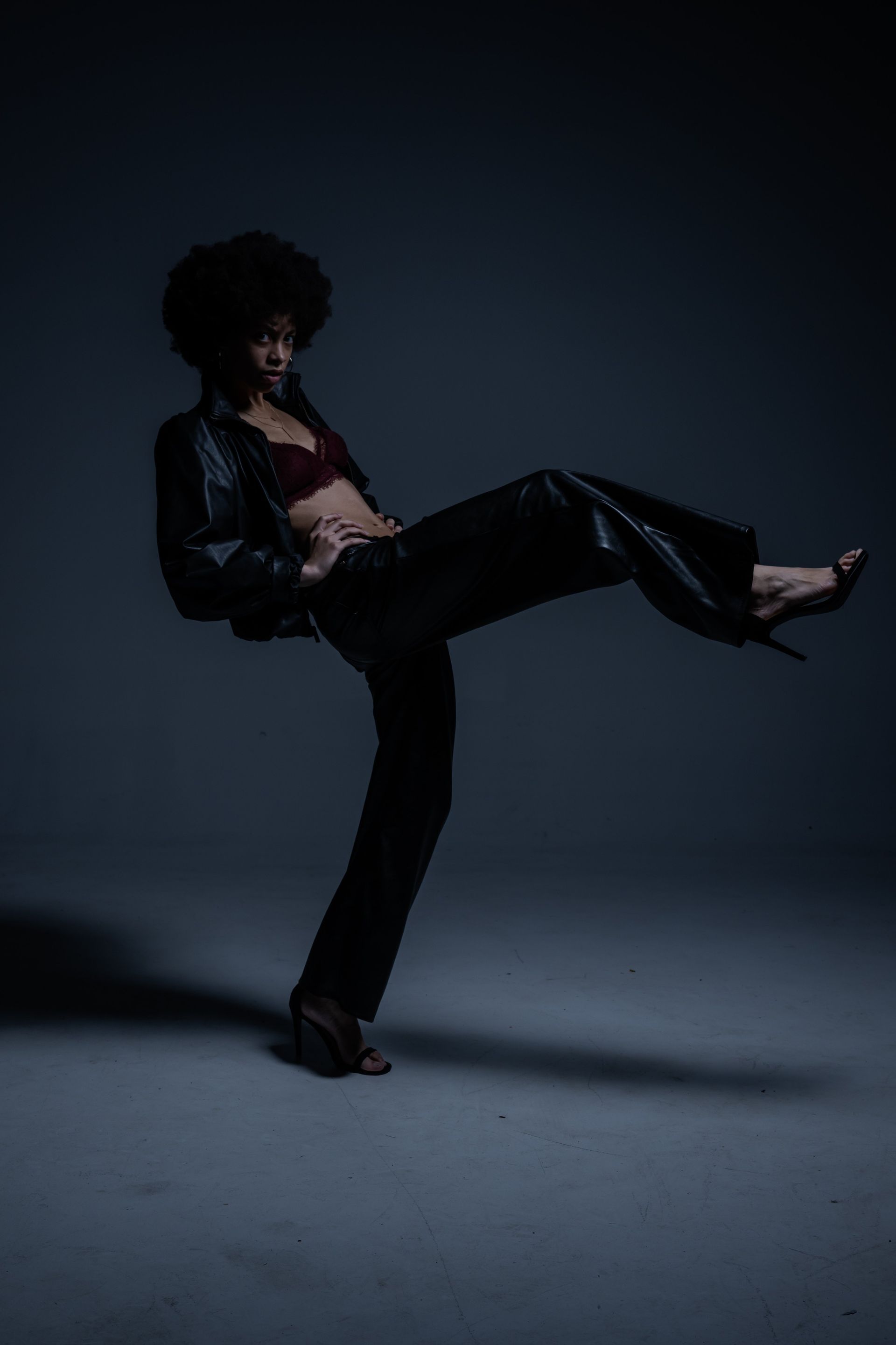 Person in black clothing posing against a dark studio backdrop, one leg lifted high.