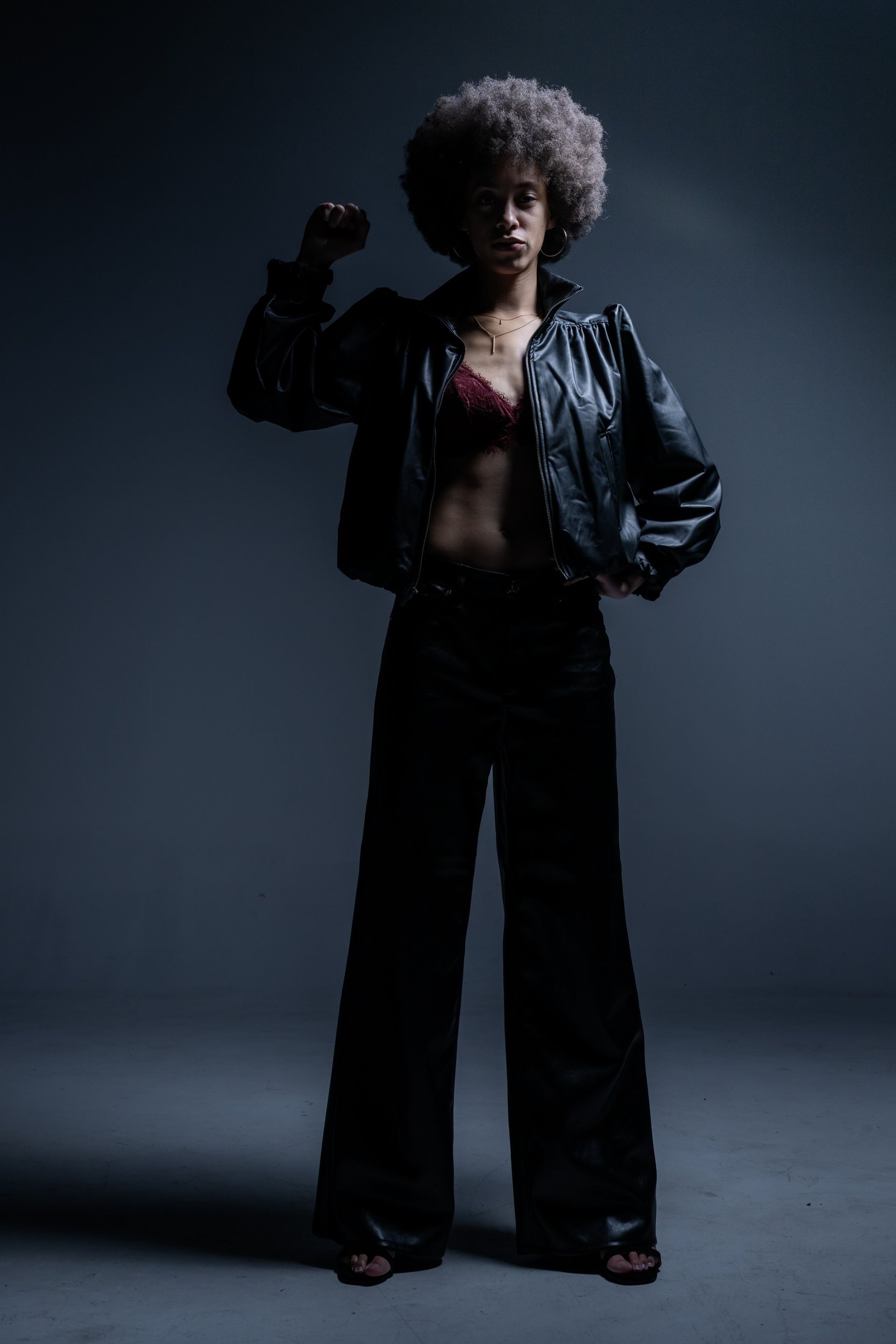 Person with afro in black jacket and wide-leg pants poses against a dark studio backdrop.
