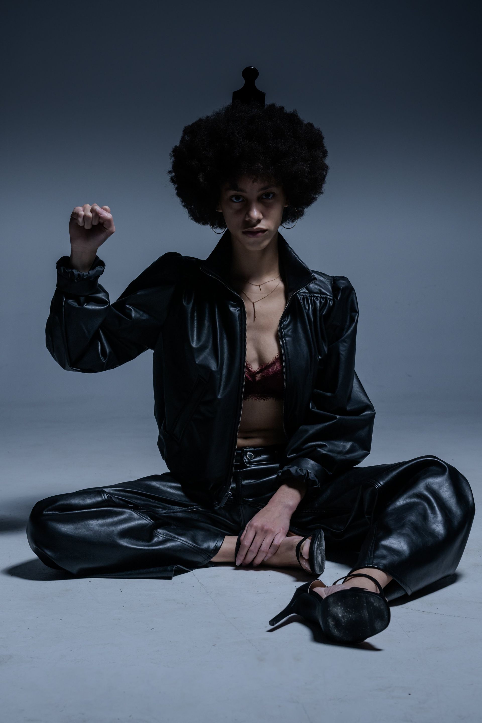 Person seated cross-legged in black leather outfit, raising a fist against a dark studio backdrop