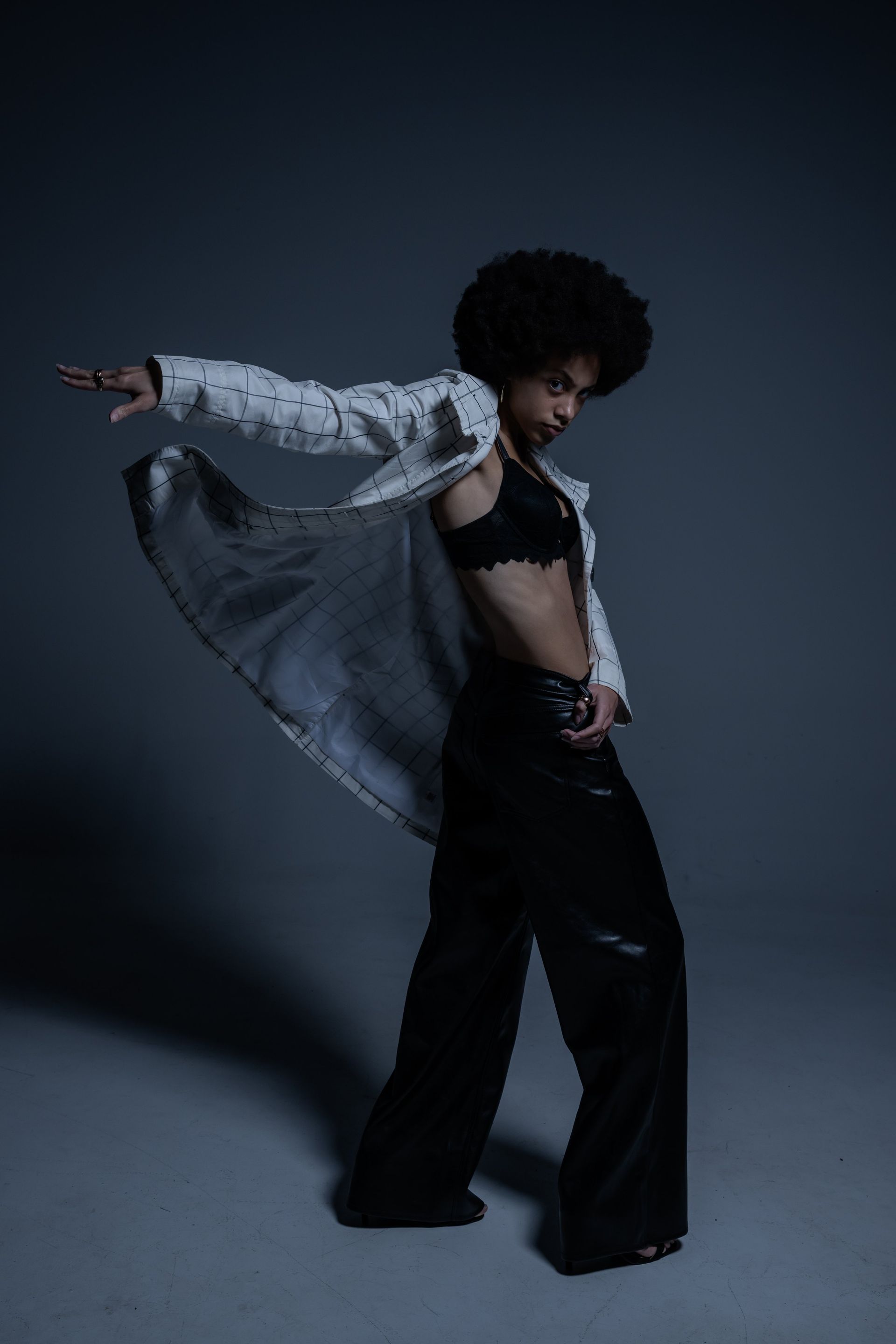 Person posing in a flowing light jacket and black pants against a dark studio background