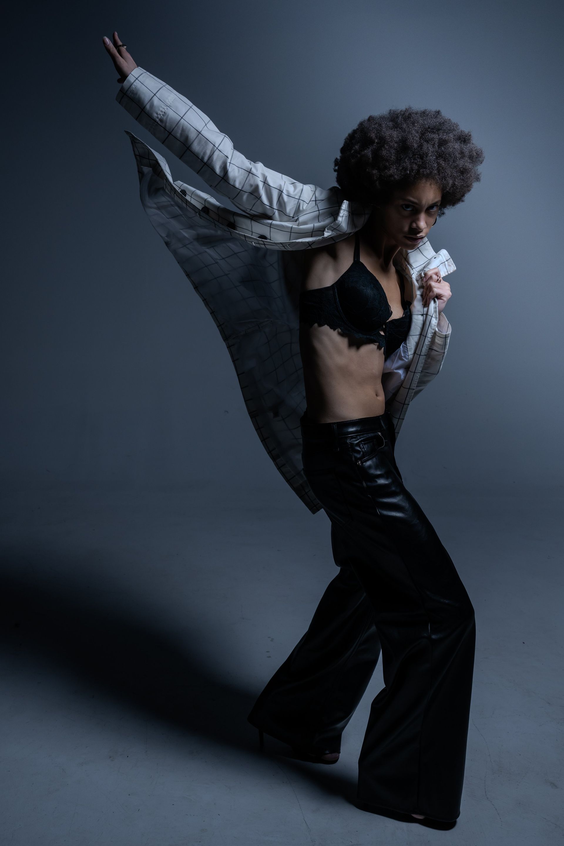 Fashion model posing in a dark studio, wearing a black outfit and silver jacket with one arm raised