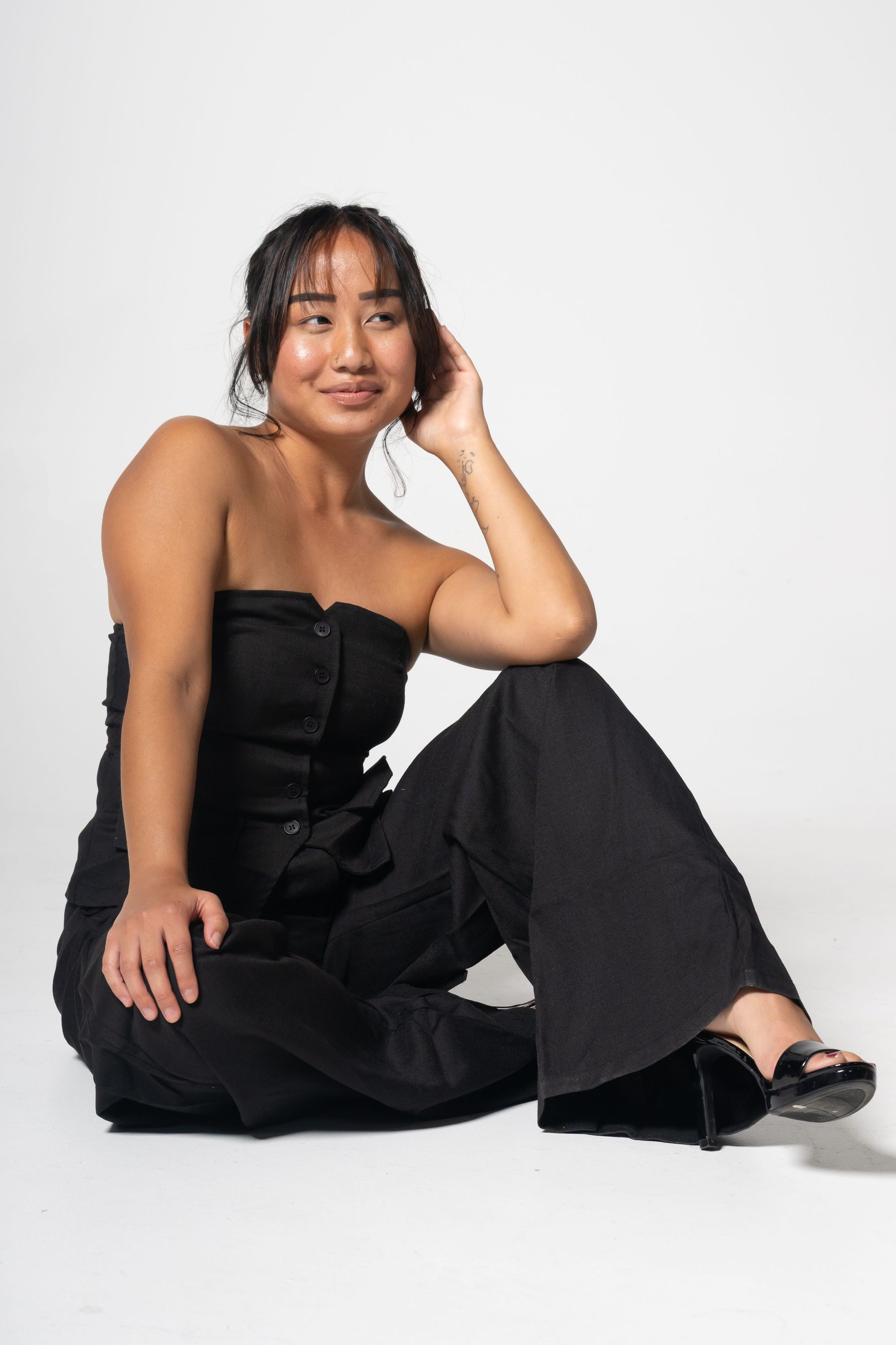 Woman in a black strapless top and pants sitting on the floor against a white background, posing with one hand on her head