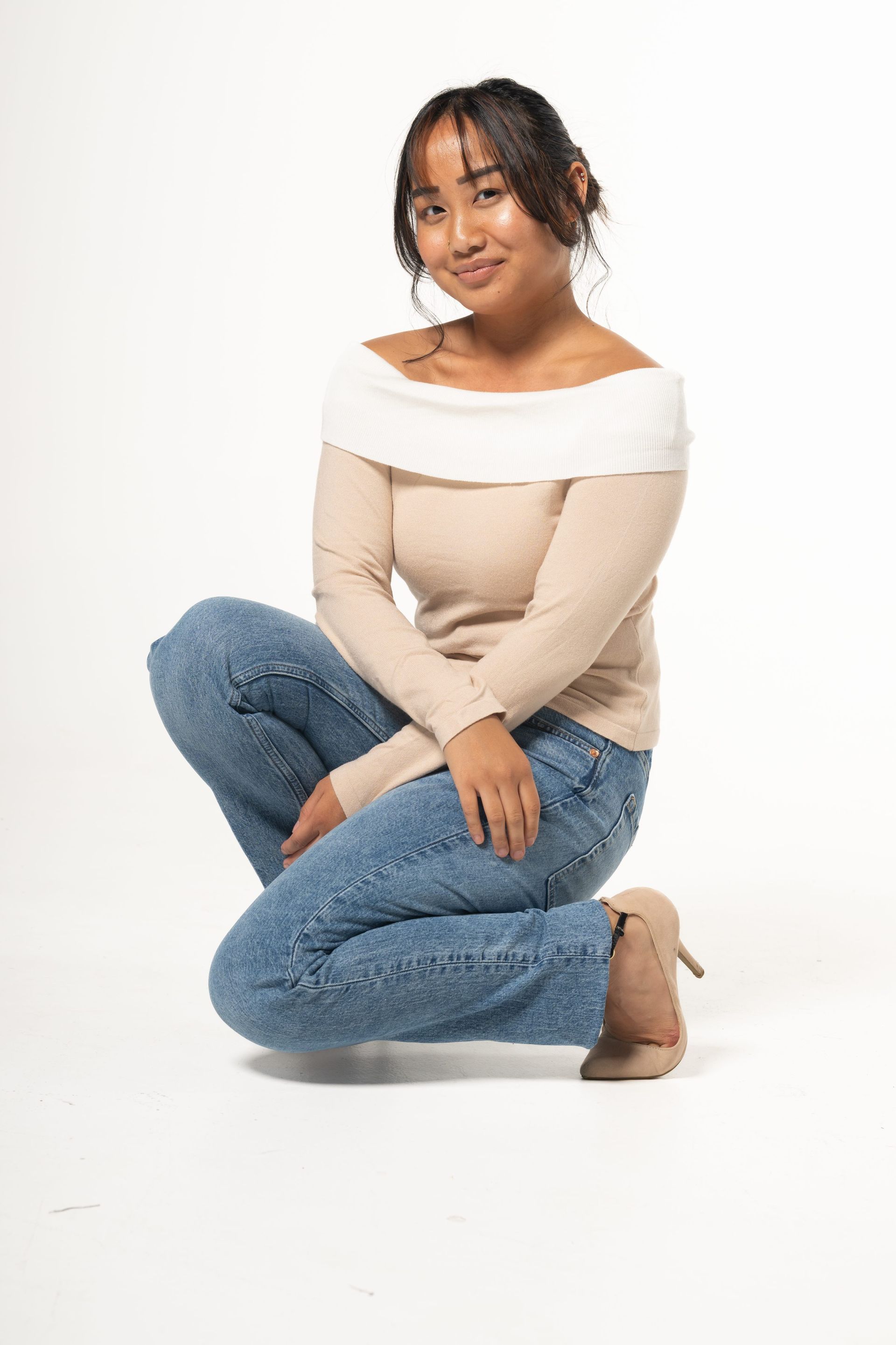 Woman crouching on a white background, wearing a white off-shoulder top and blue jeans.