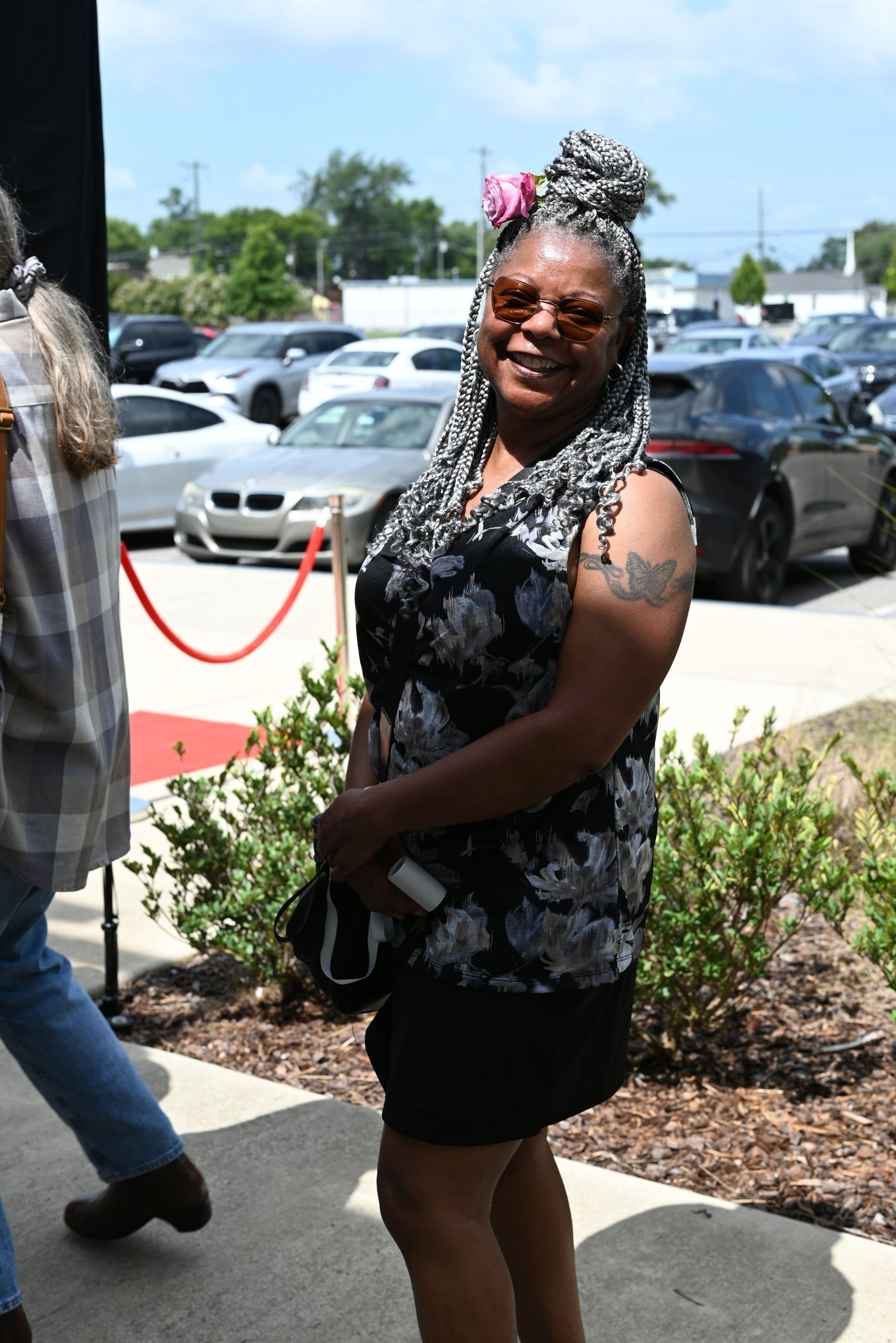 Woman in black floral top and shorts smiling outdoors near parked cars, with a red rope barrier nearby