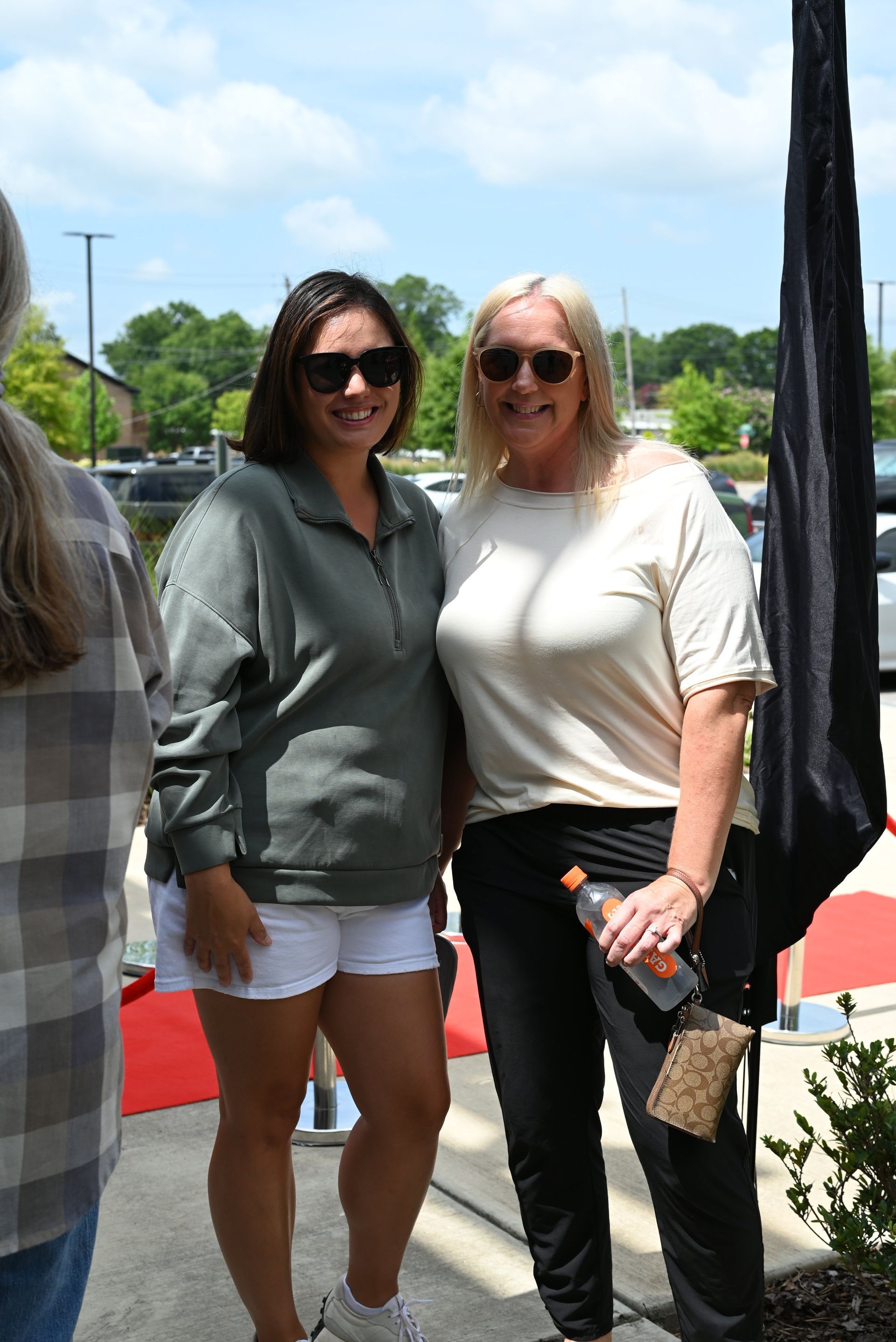Two women smiling outdoors, standing on a red carpet in bright daylight.