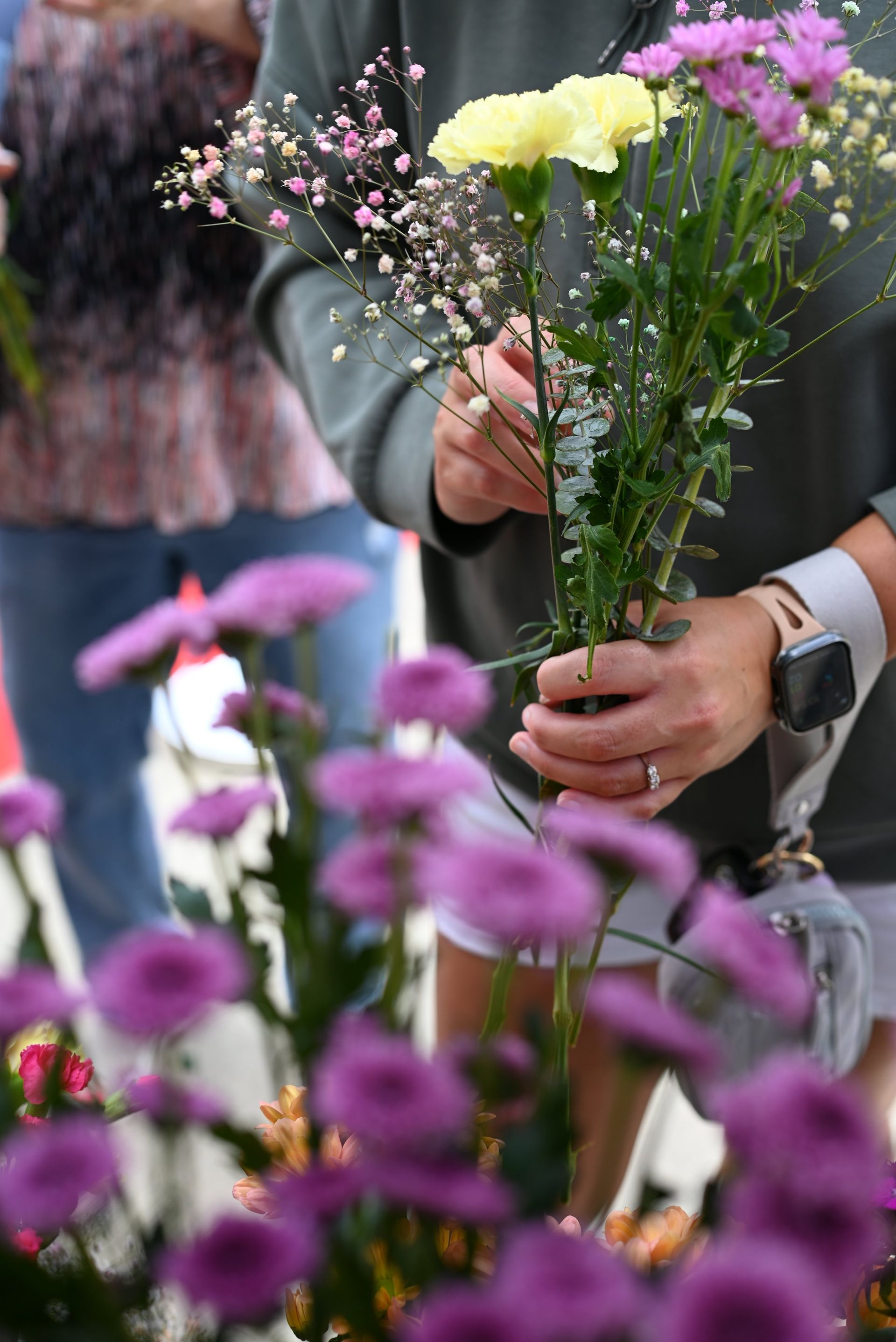Hands arranging pastel flowers at an outdoor market, with purple blooms in the foreground.