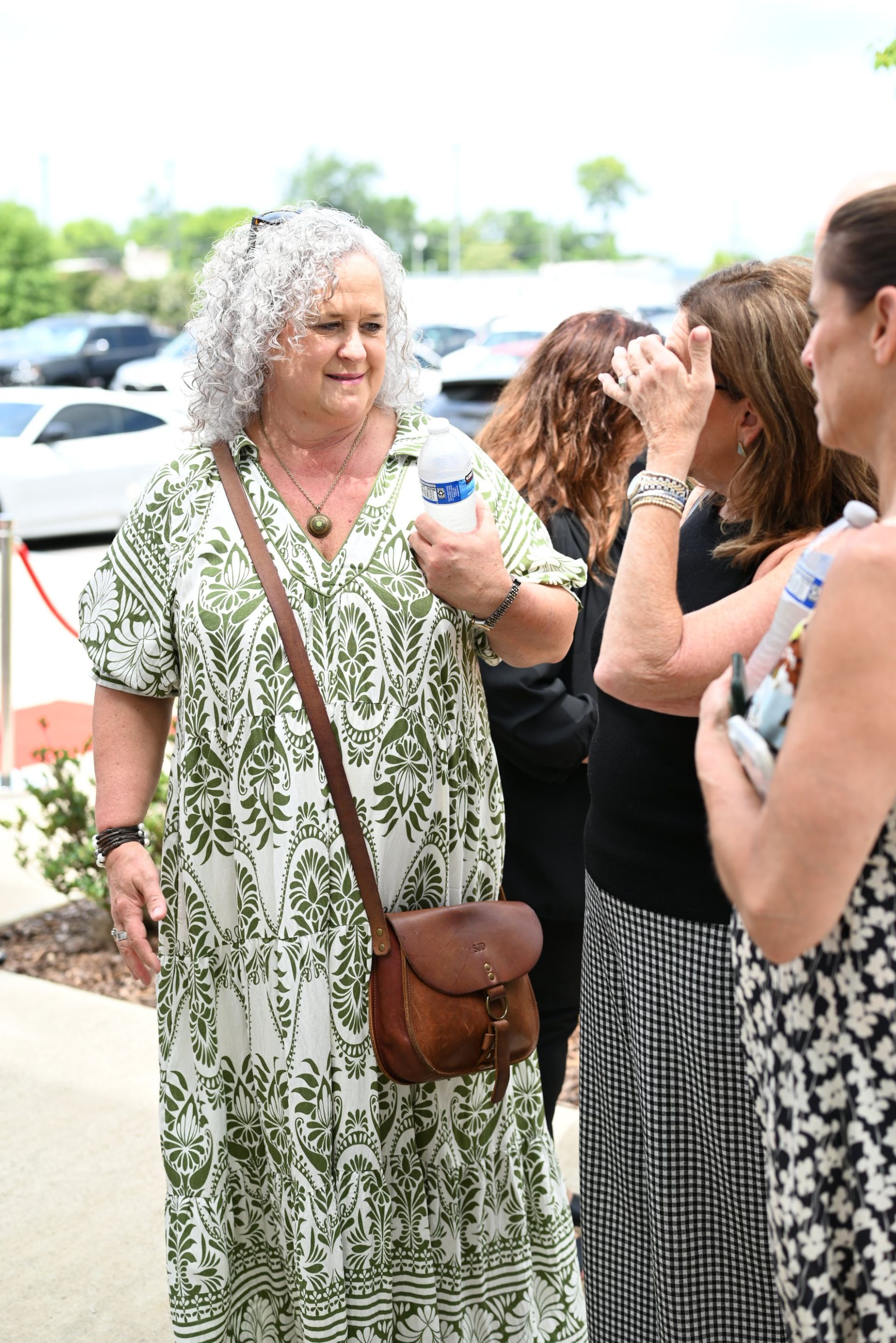 Woman in green patterned dress with cup, talking with others outdoors near parked cars.