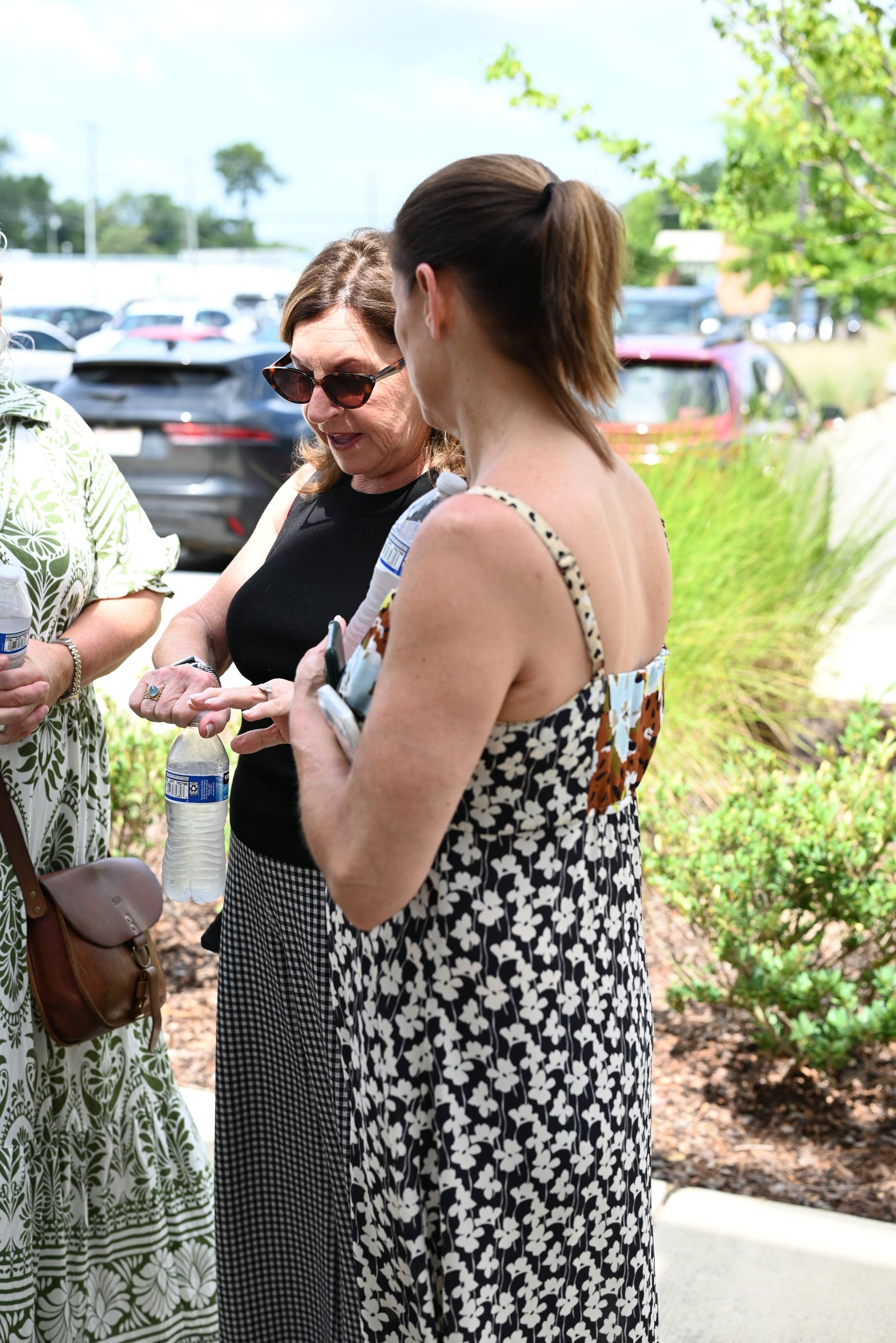 Two women chatting outdoors near parked cars, one in a black floral dress, the other in sunglasses.