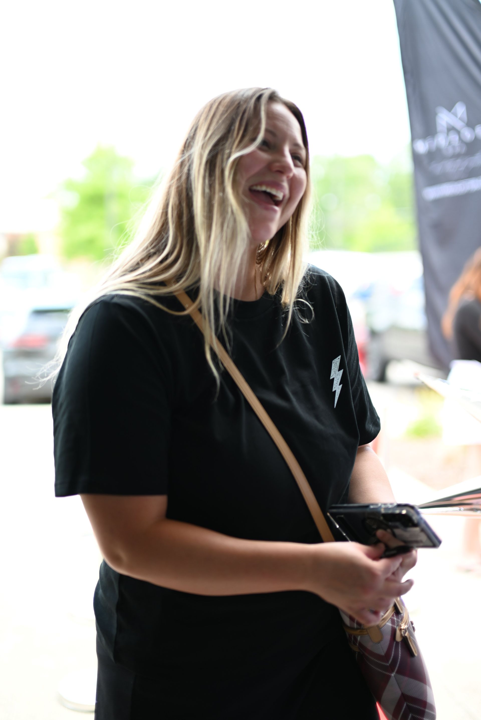 Person smiling while holding a camera, wearing a black T-shirt and crossbody bag indoors.