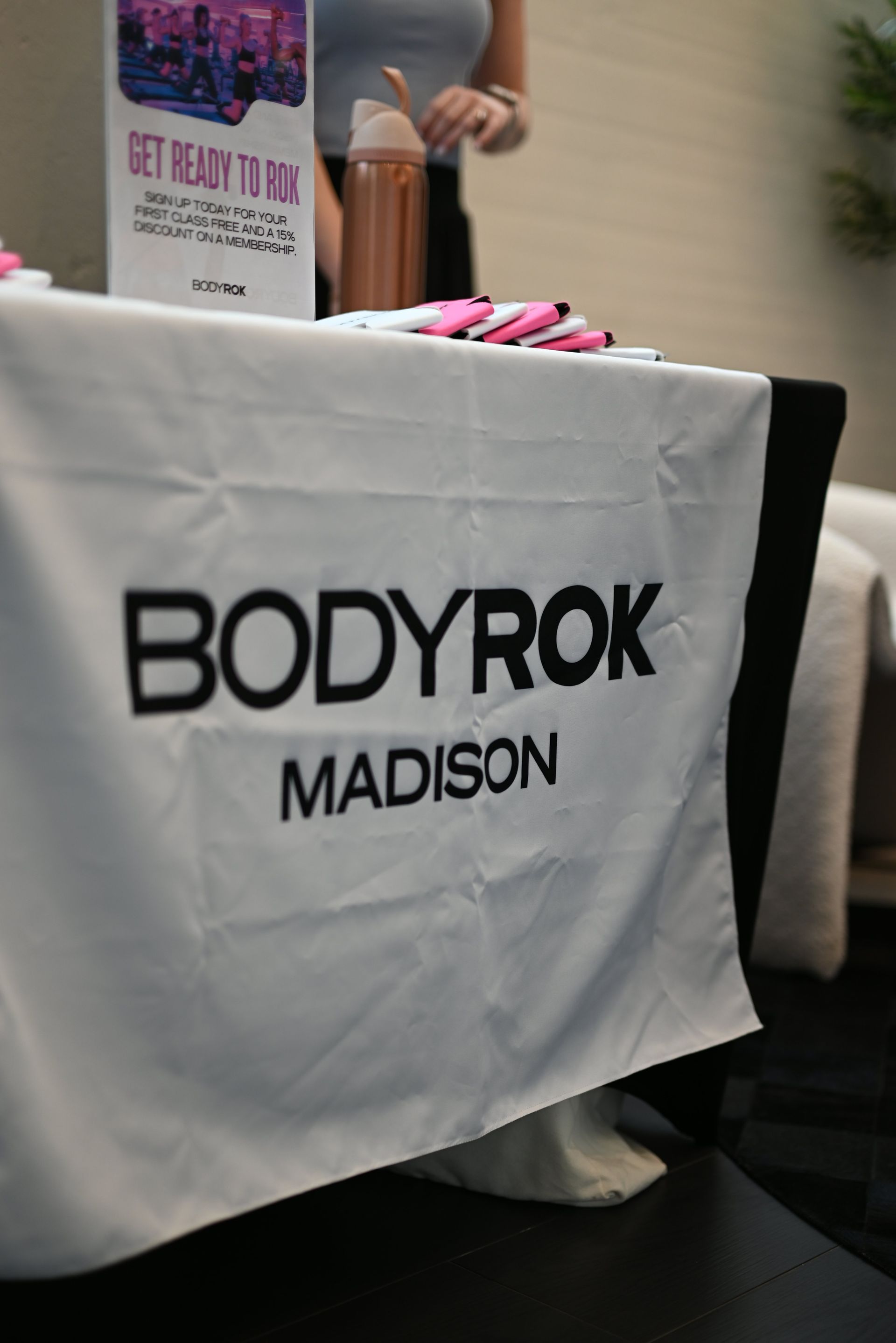 Bodyrok Madison sign on a white tablecloth at an indoor event, with pink items and a drink behind it