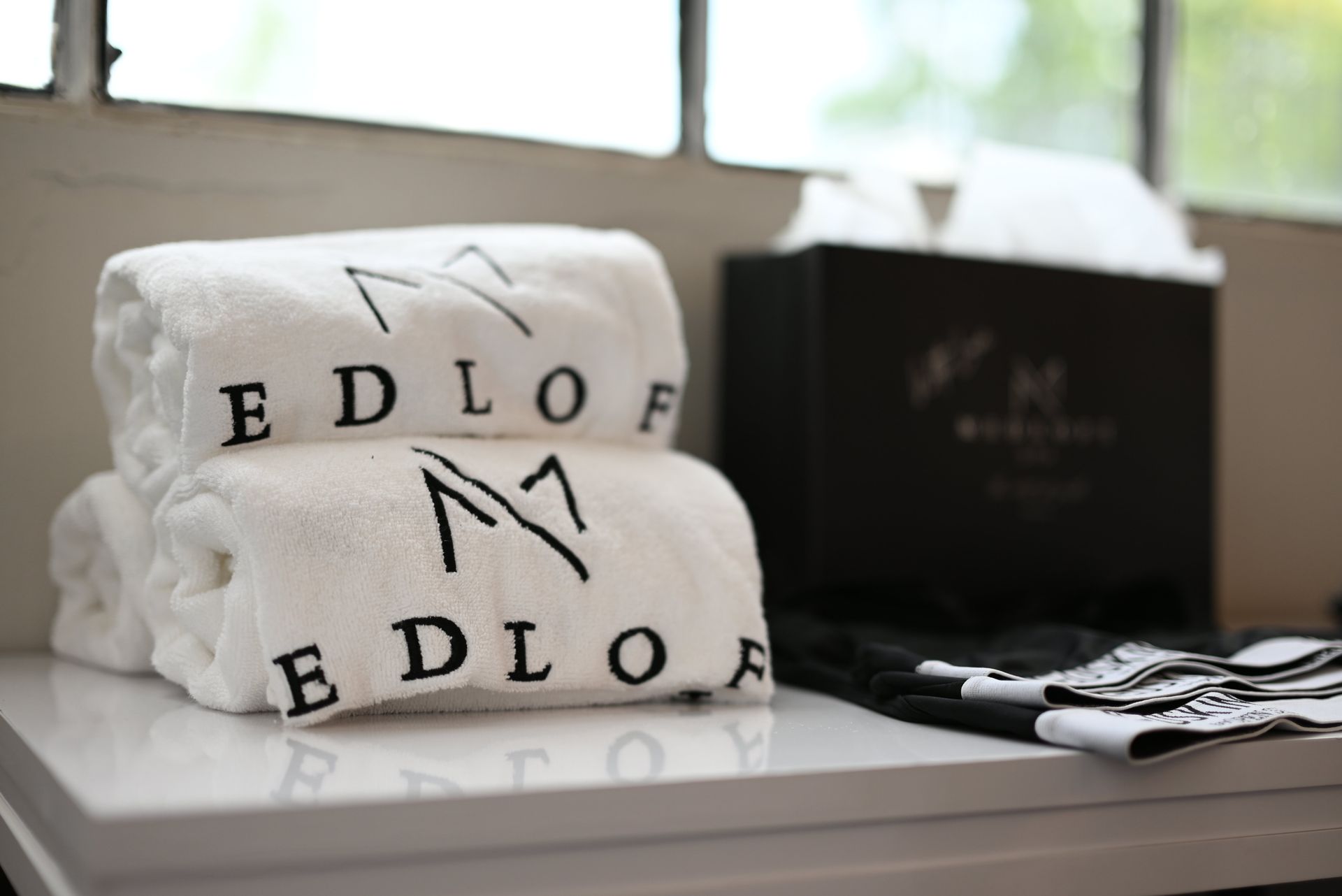 Folded white towels with black “EDLOF” lettering on a bathroom counter beside toiletries.