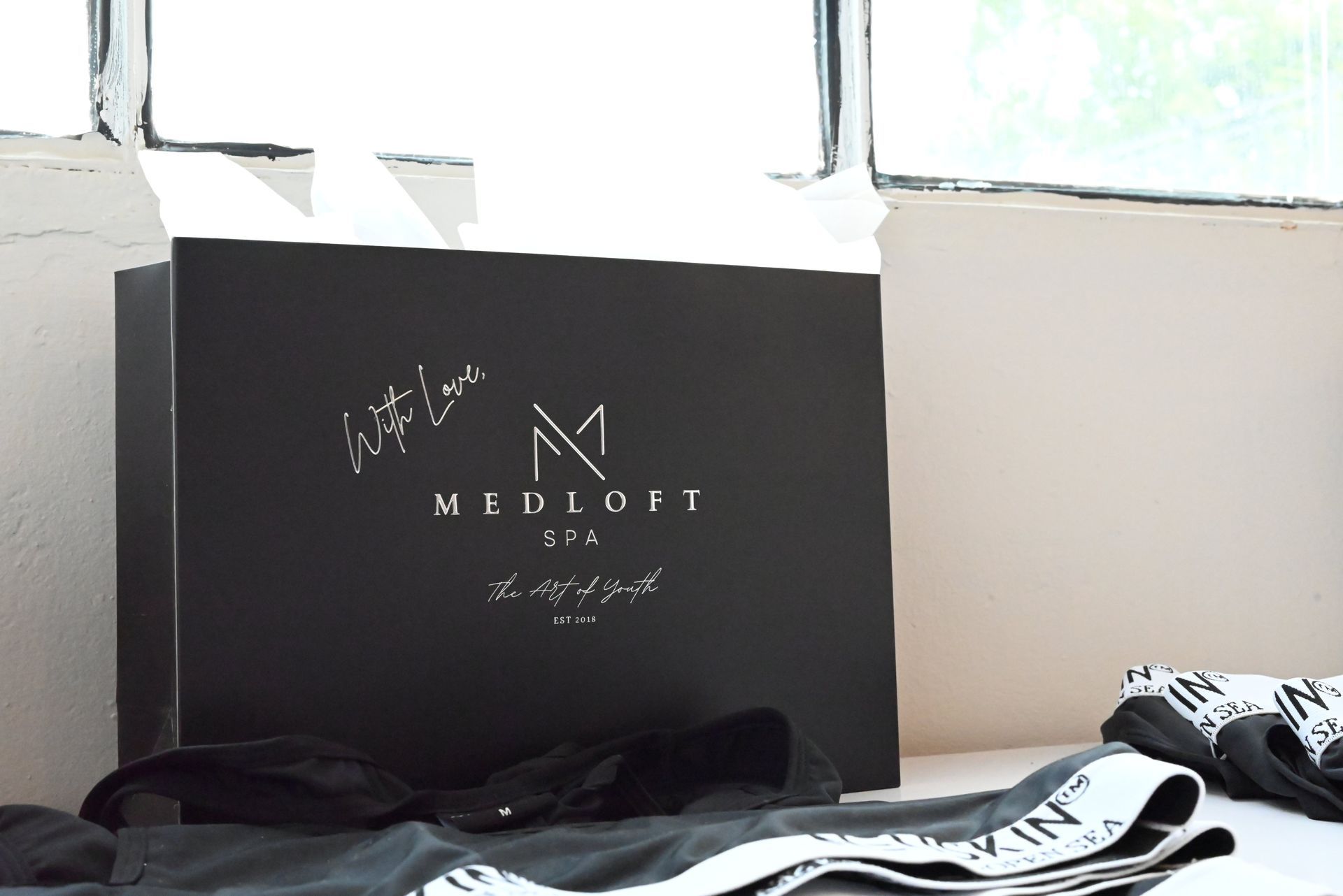 Black shopping bag with white logo on a bed near a window, with folded clothes in the foreground