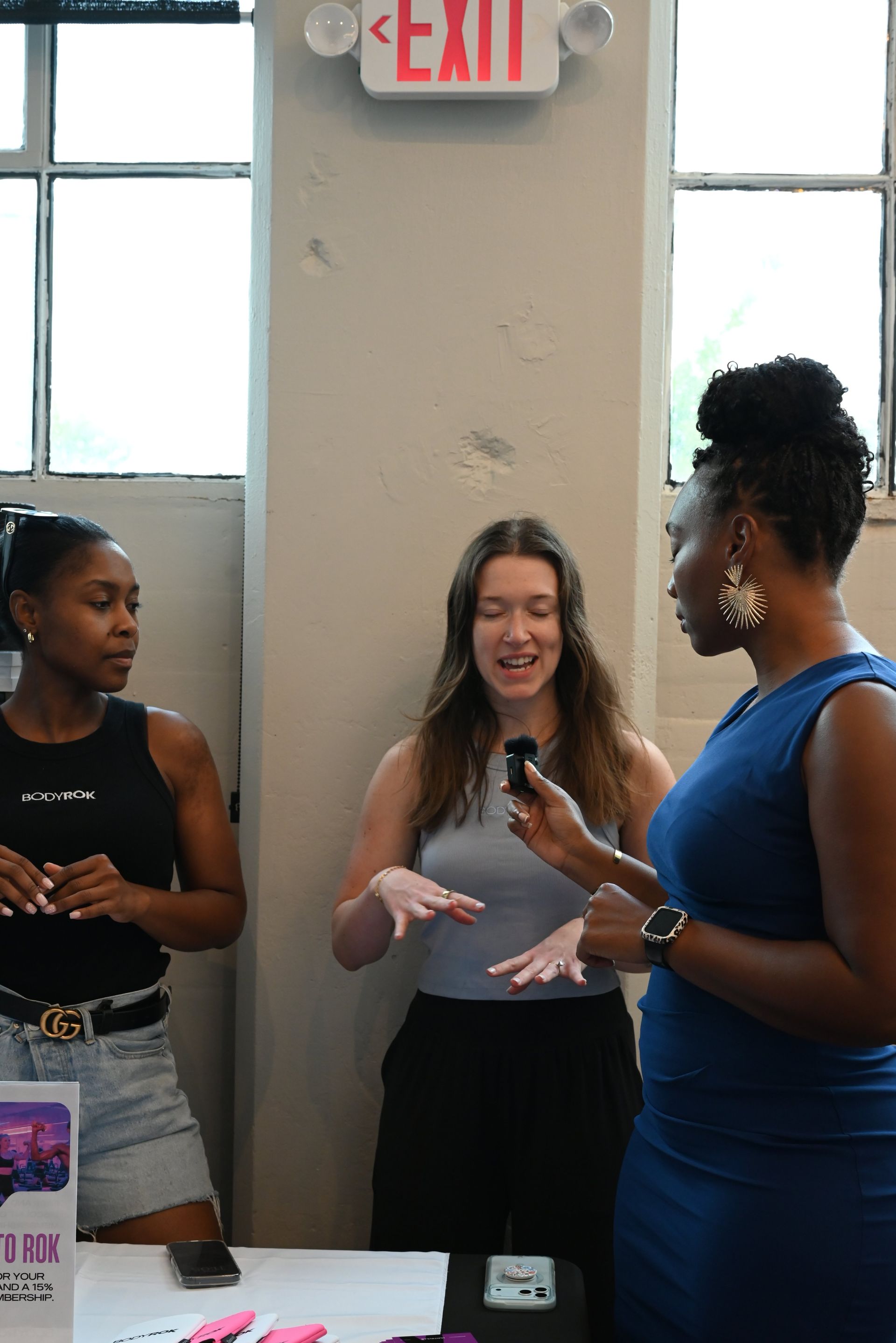 Three women chatting at a table in a bright indoor event space near a window and exit sign