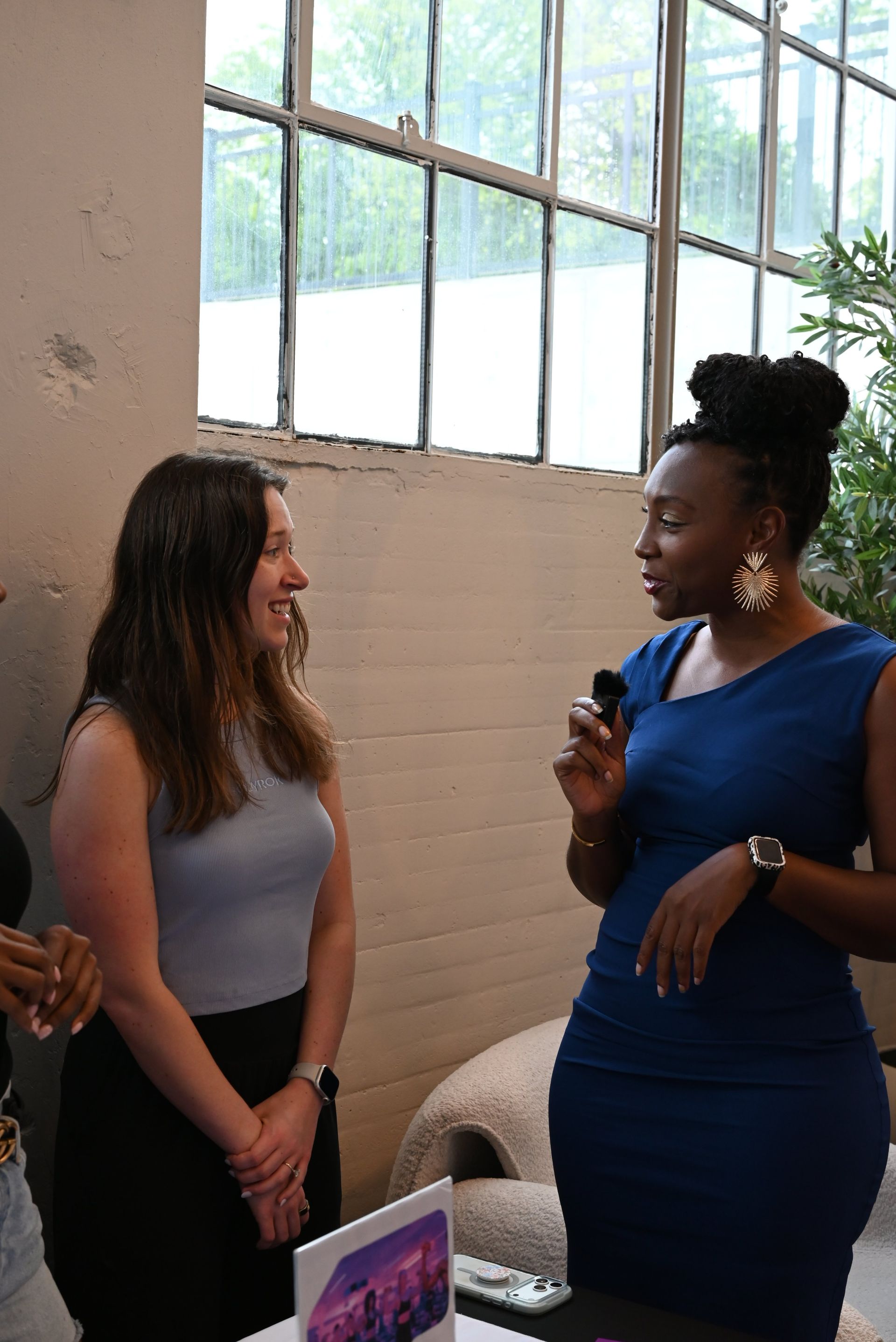 Two women chatting indoors by a window, one holding a microphone and wearing a blue dress.