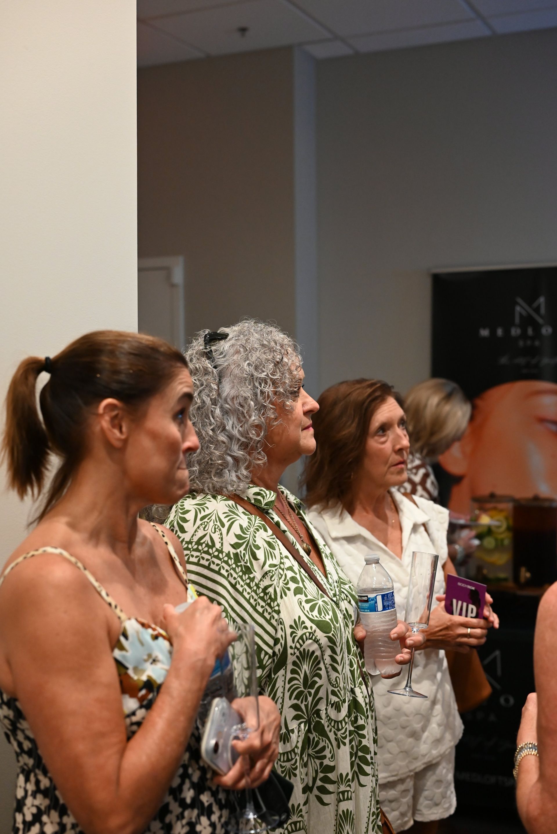 Four women standing indoors at an event, holding drinks and looking toward the right.
