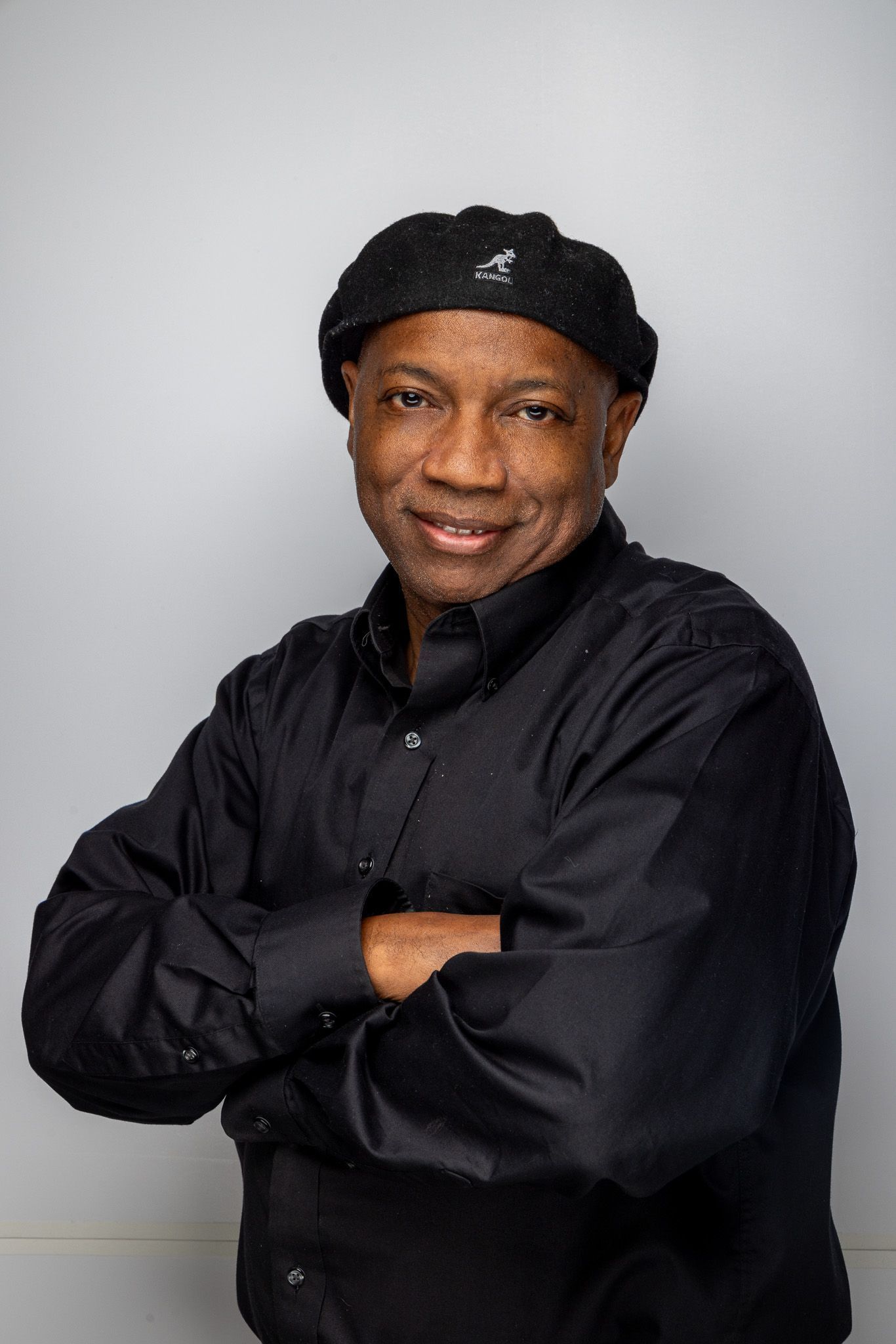 Smiling man in black shirt and cap, standing with arms crossed against a gray background