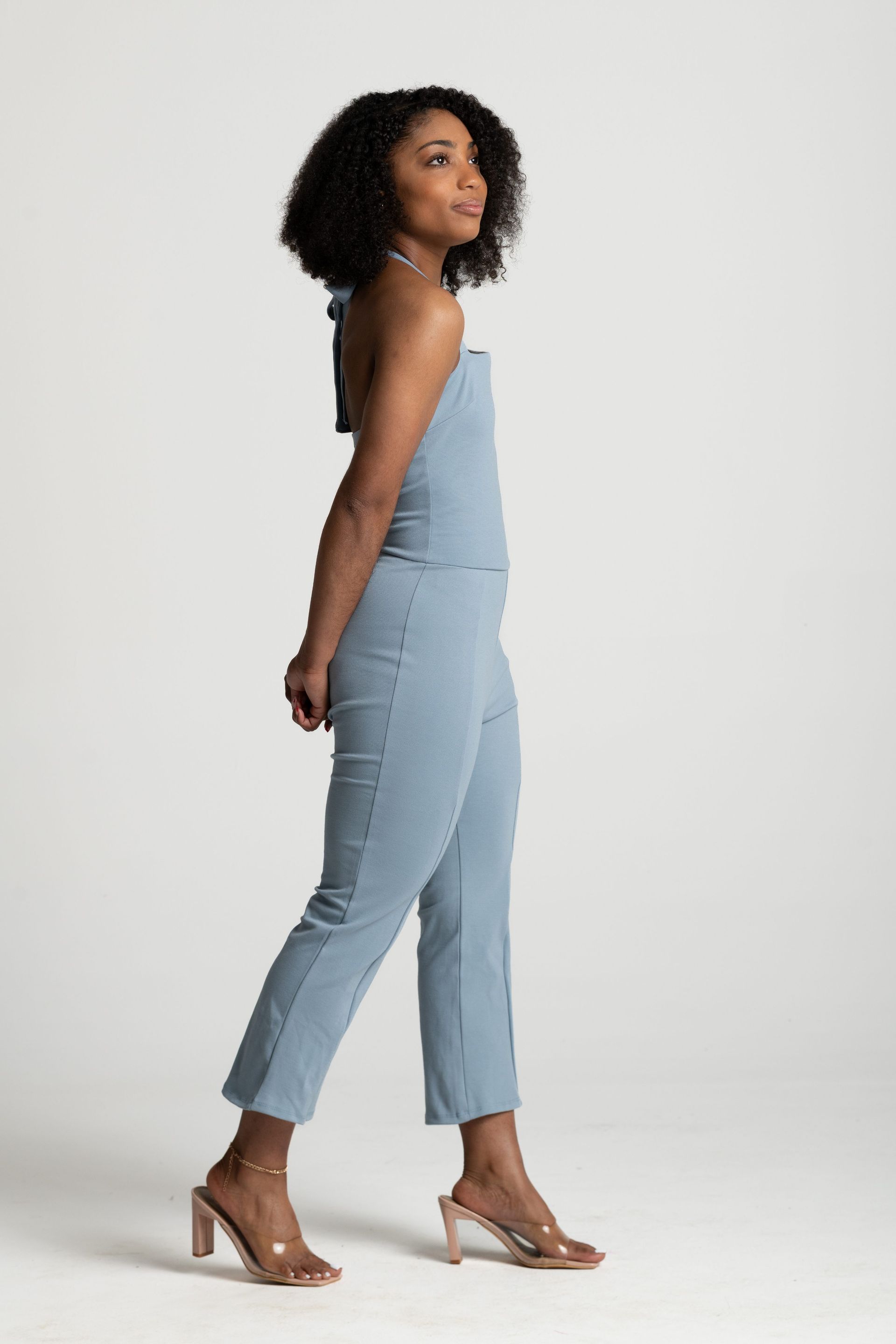 Woman in a light blue jumpsuit and heels posing in profile against a plain white background