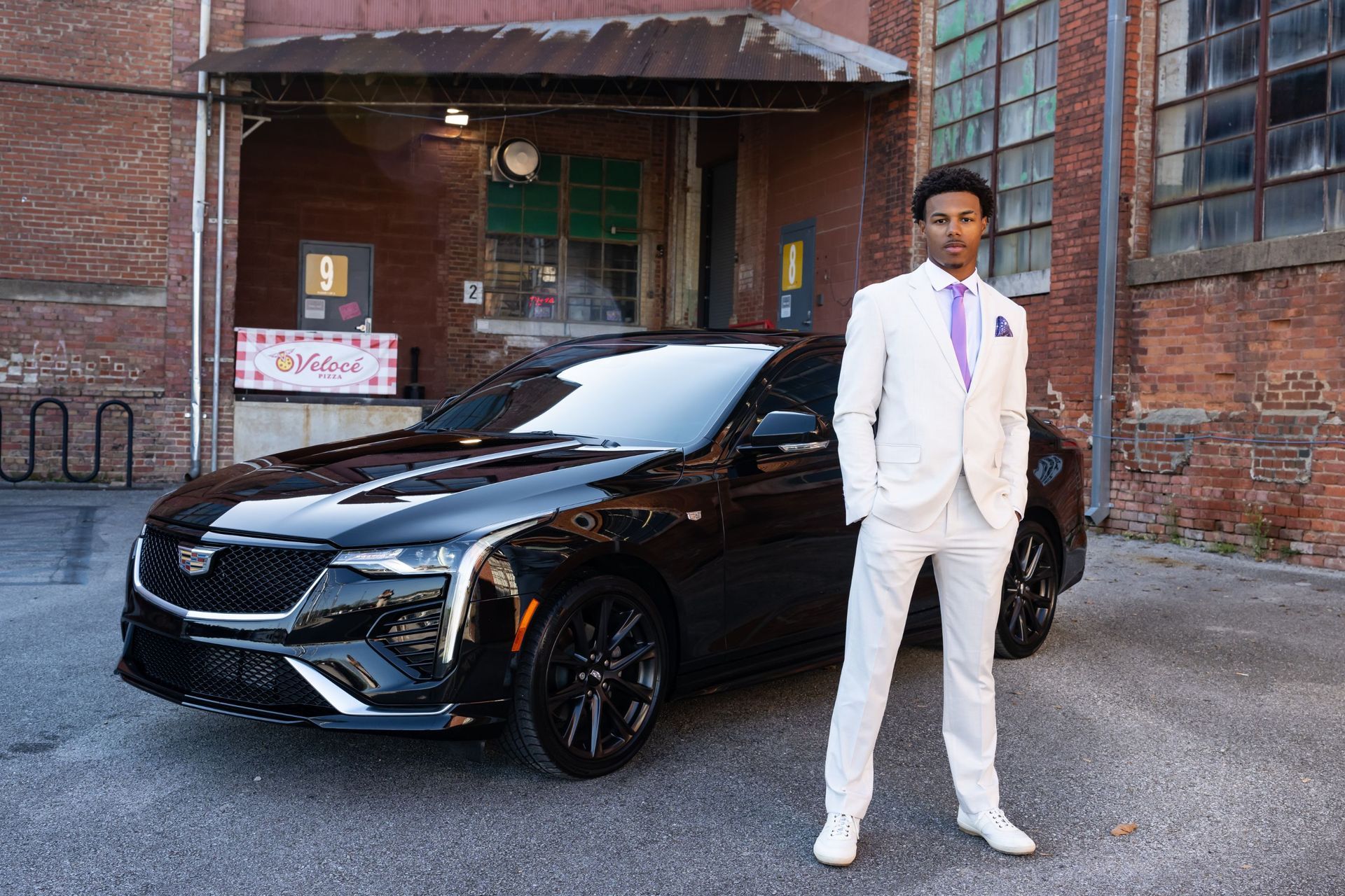 Man in a white suit standing beside a black sedan outside a brick building