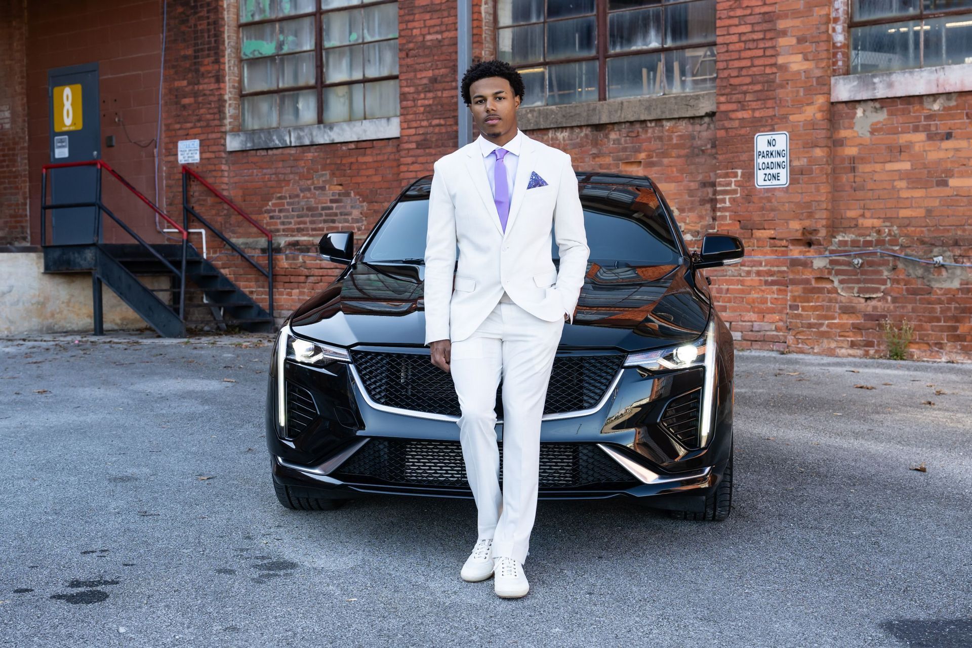 Man in a white suit standing in front of a black sports car outside a brick building.