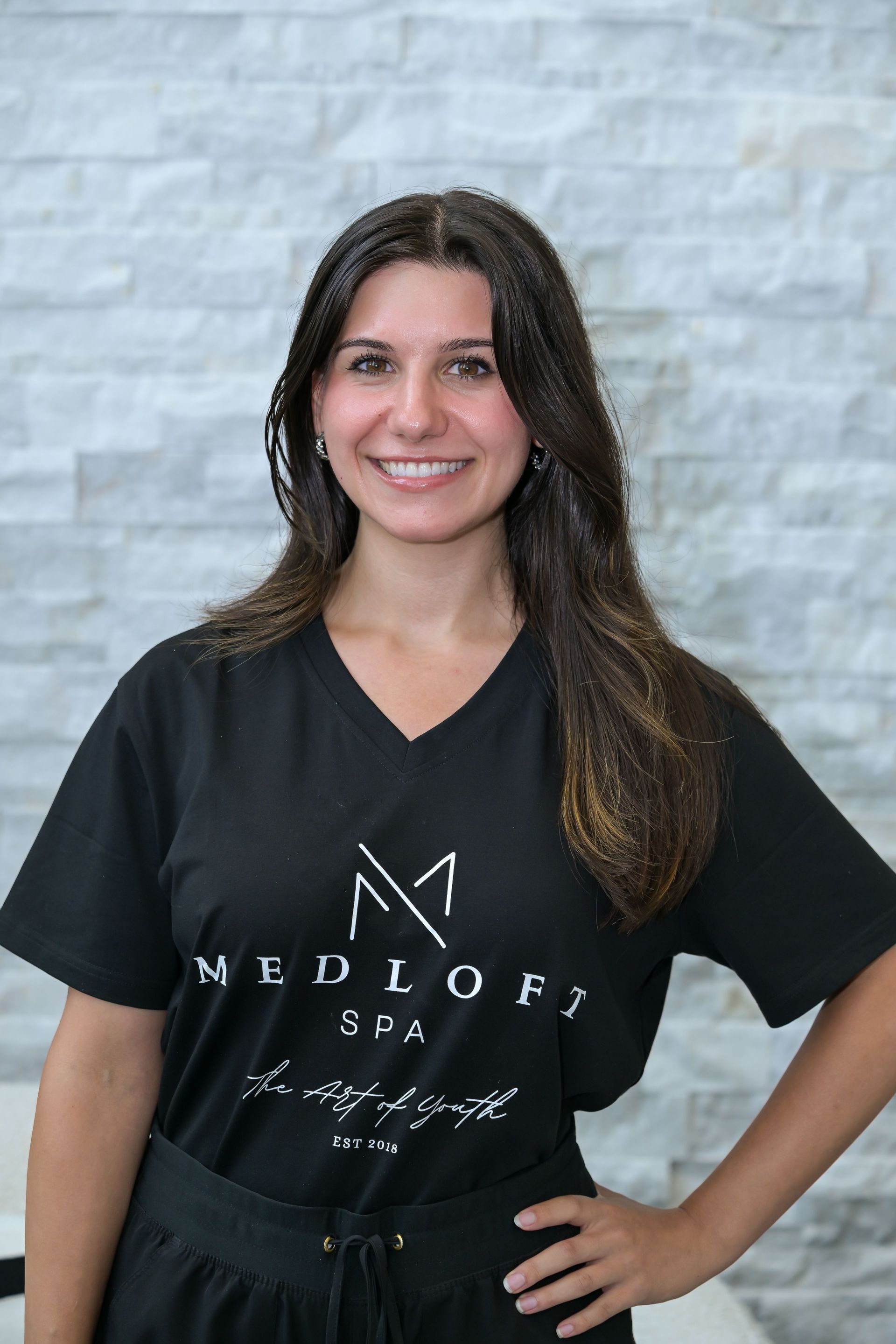 Smiling person in a black MedSpa T-shirt standing with hands on hips against a light brick wall