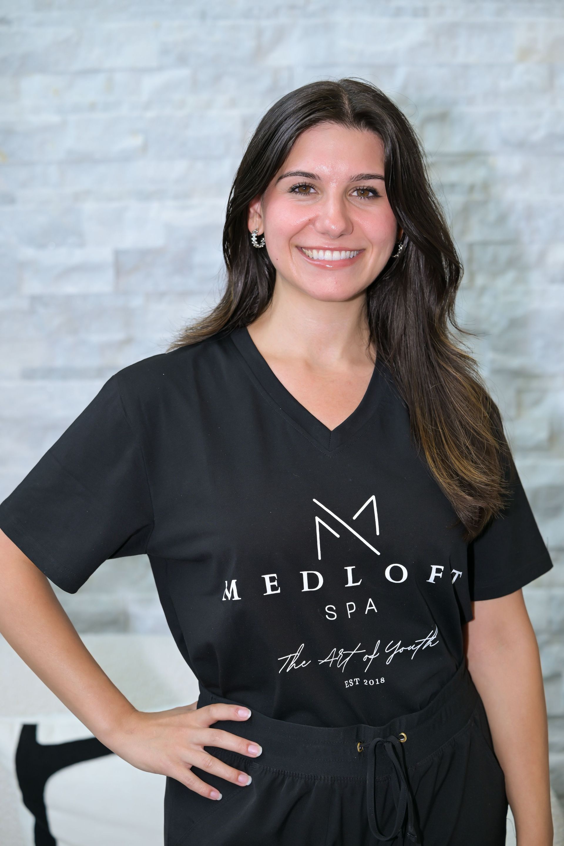 Smiling person in black scrubs with hand on hip against a light gray backdrop