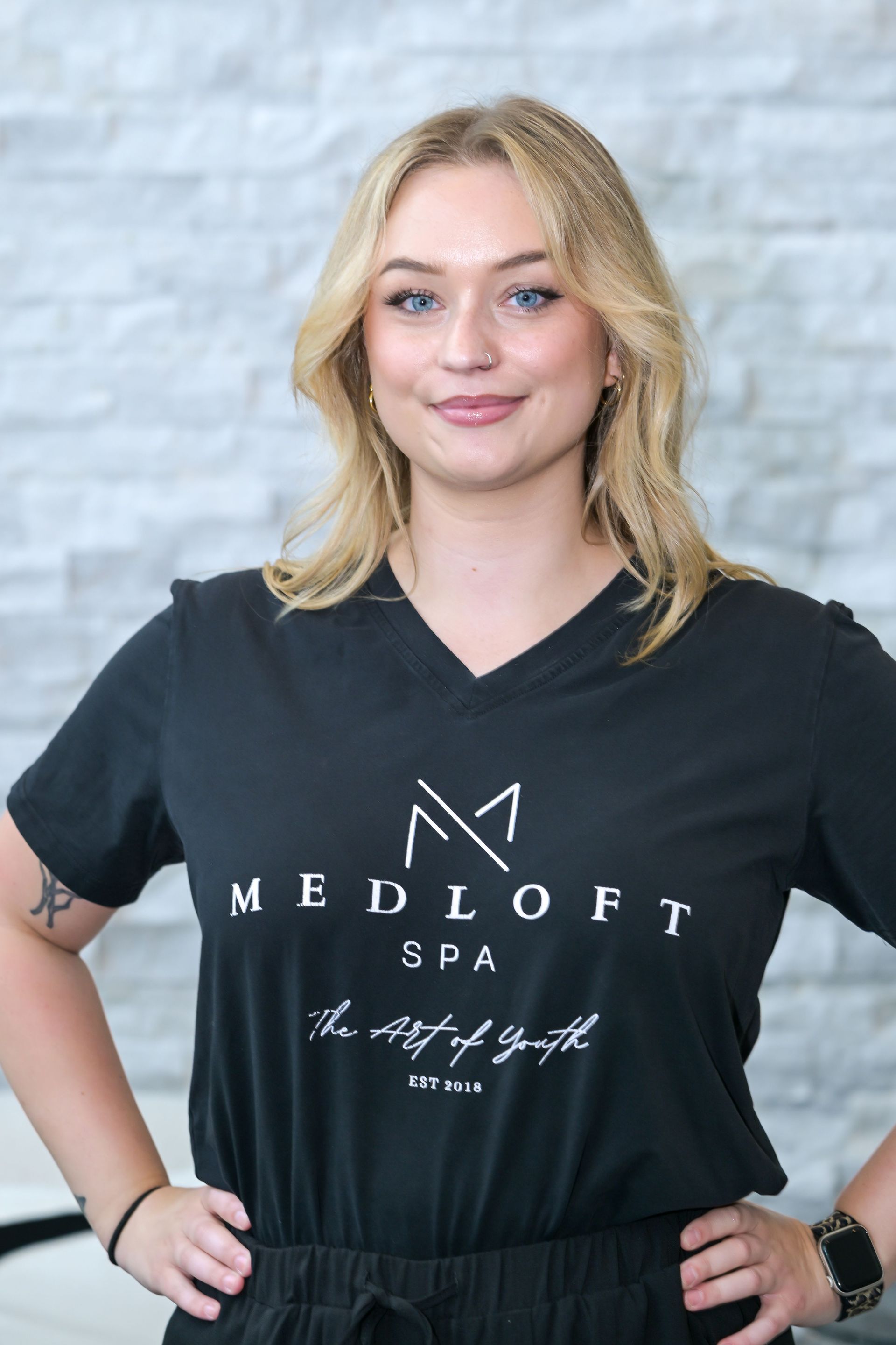 Woman in black Medloft Spa T-shirt and skirt, standing with hands on hips and smiling.