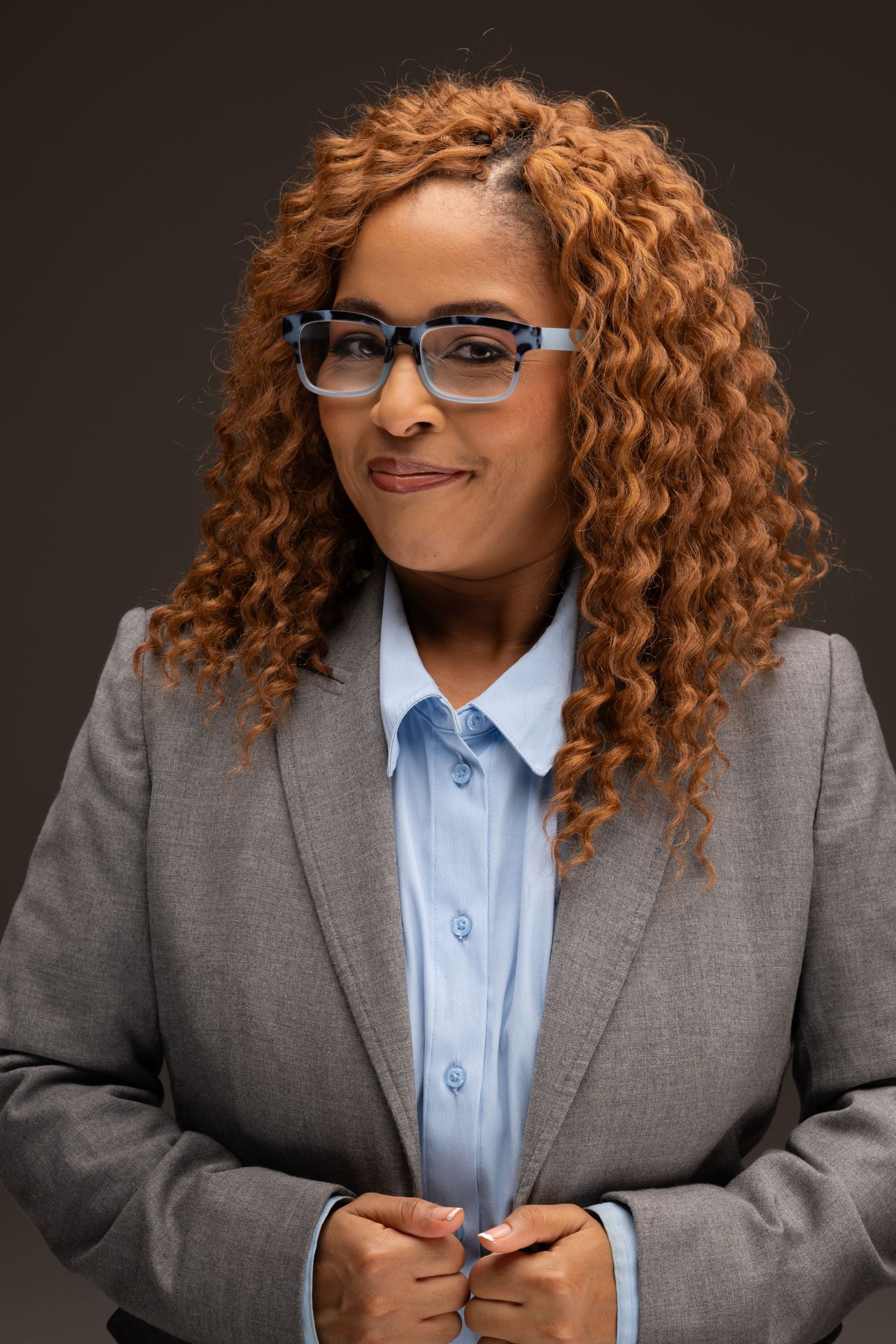 Portrait of a person in a gray blazer and blue shirt, wearing glasses, standing against a dark background
