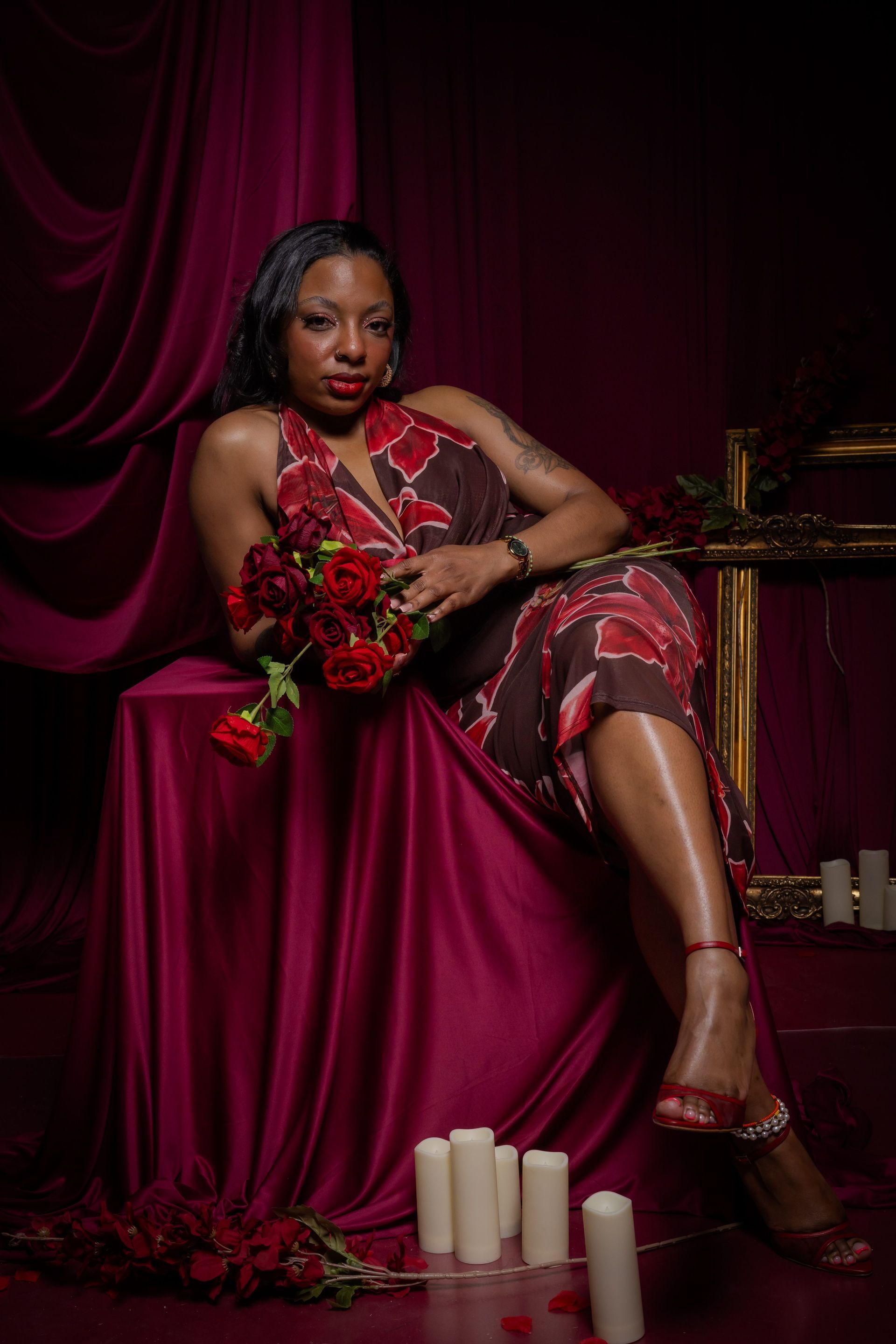 Woman in a burgundy gown seated by candles and flowers against a dark draped backdrop