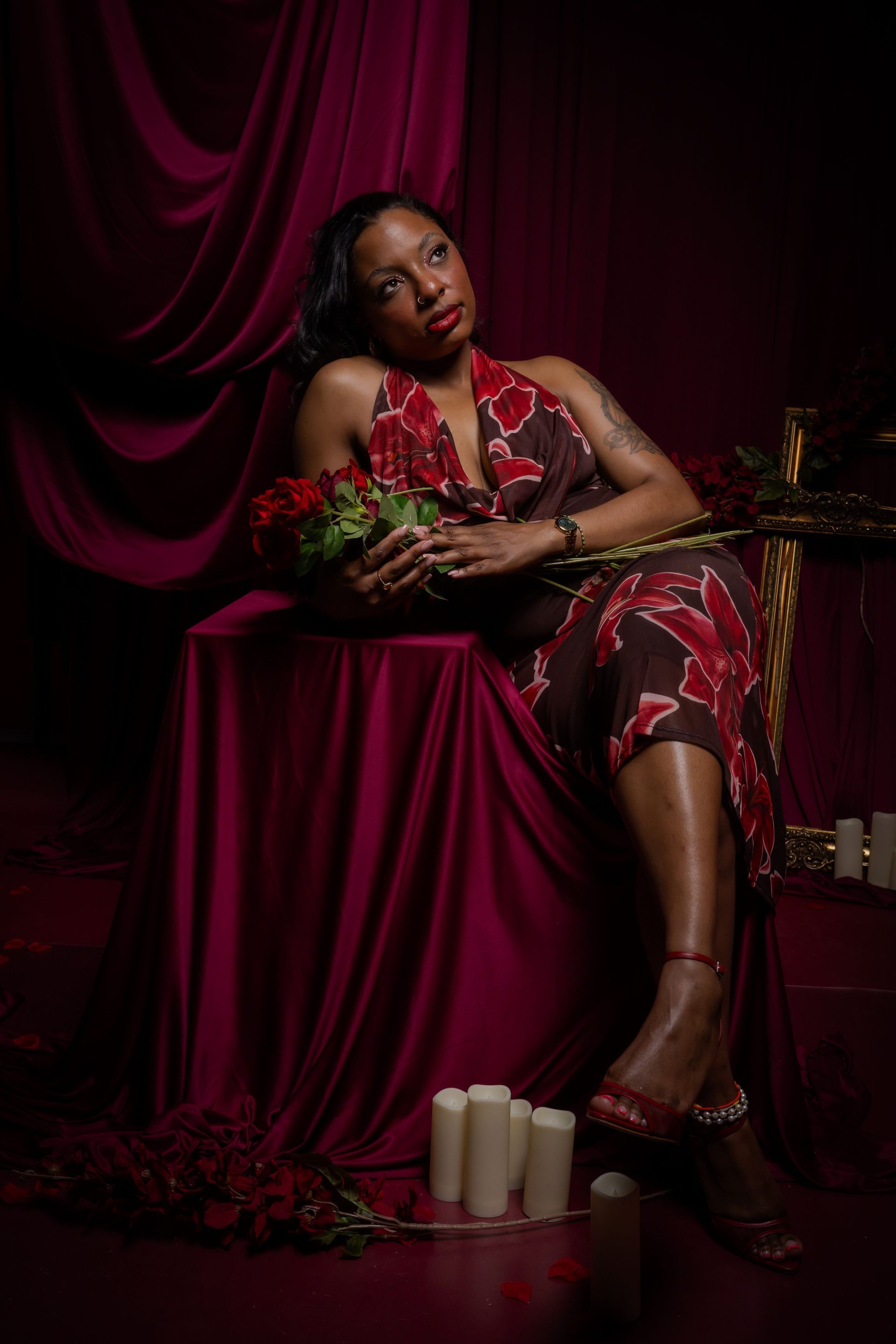 Woman in a red dress reclining on a draped chair, holding flowers in a dark studio setting