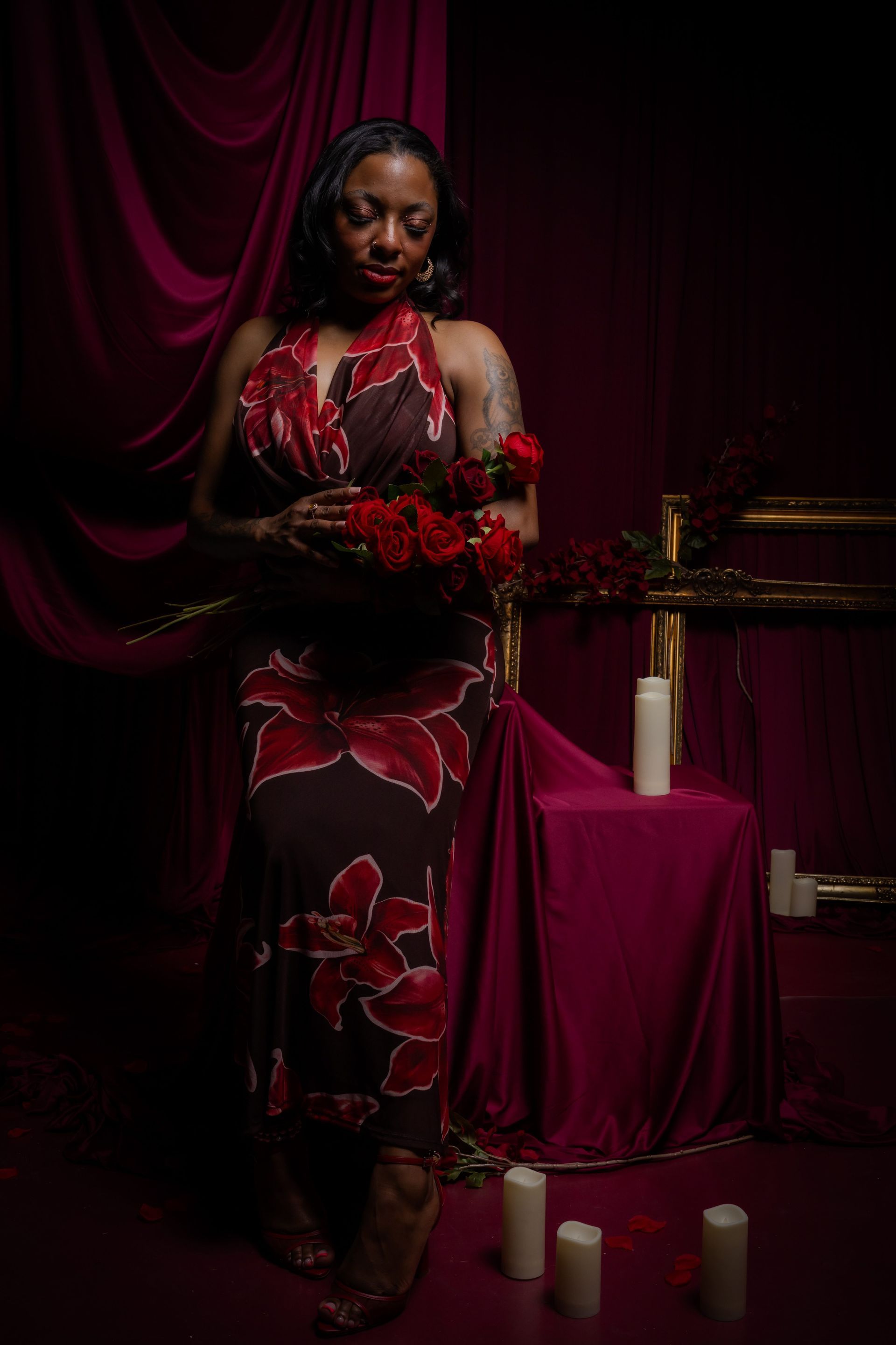 Person in a red floral dress holding red flowers in a dimly lit room with candles and burgundy drapes