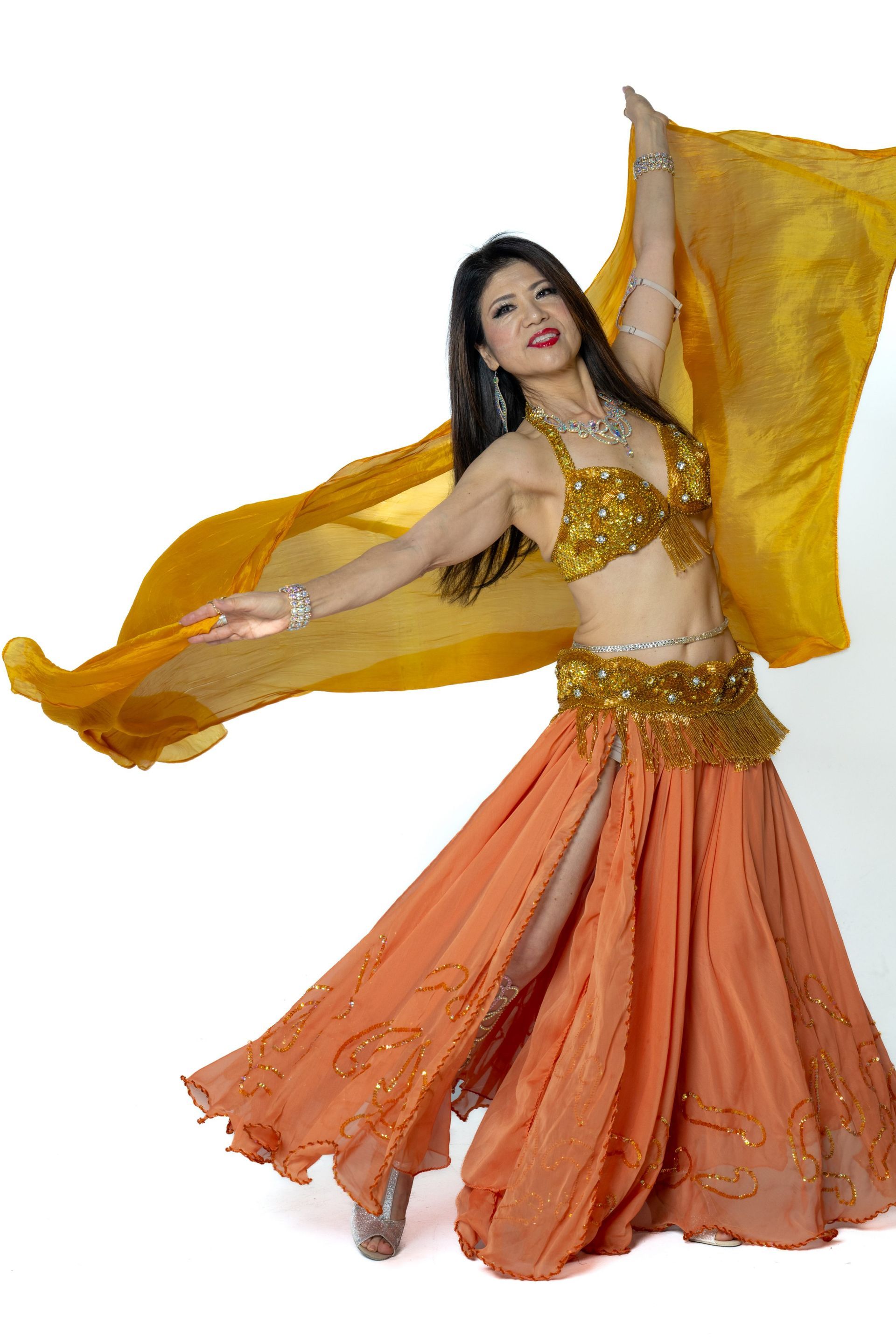 Woman in a flowing orange and gold dance costume posing with raised arms and sheer fabric against a white background