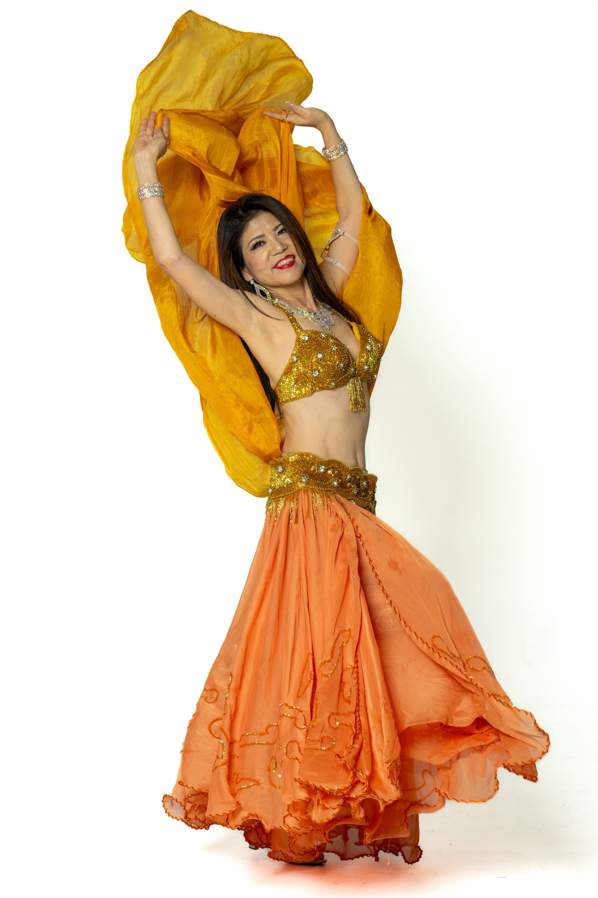 Woman in a gold top and orange skirt dancing with flowing yellow fabric against a white background