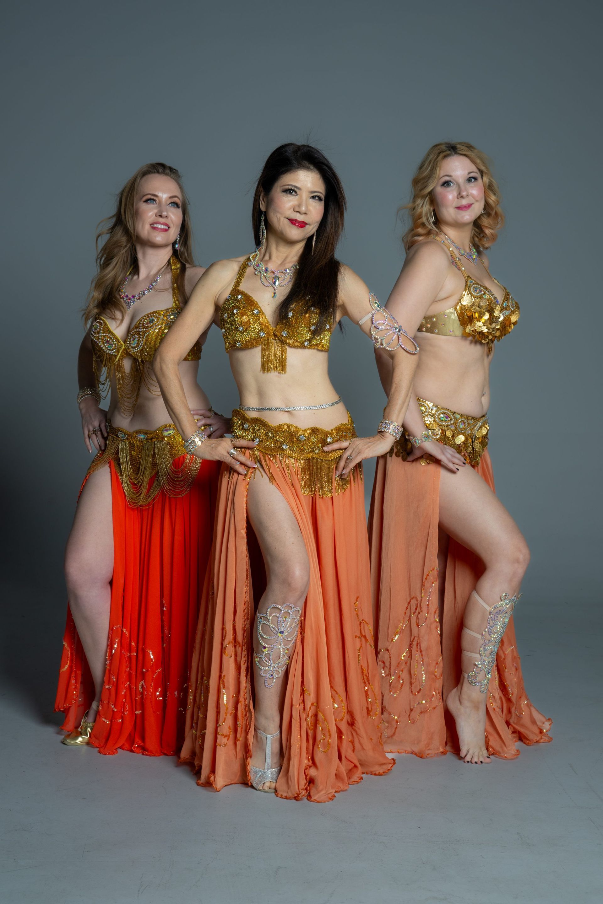 Three dancers in gold costumes with orange and red skirts, posing in a studio backdrop