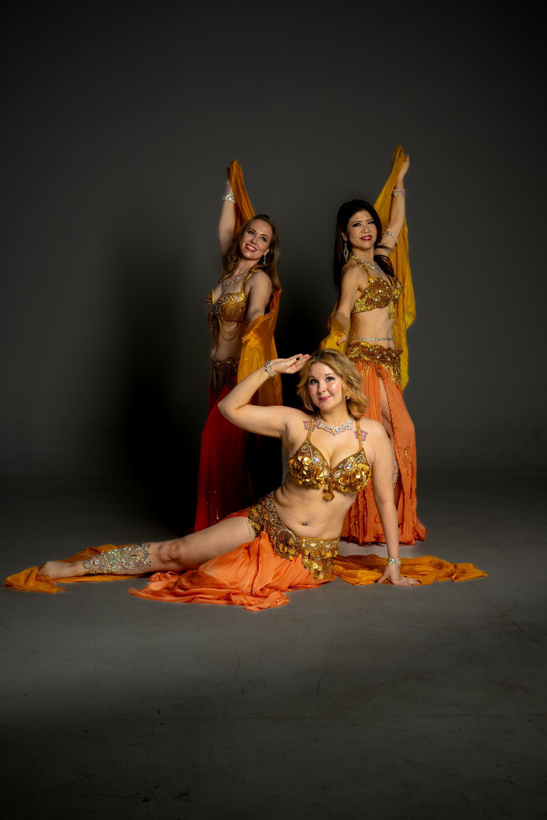 Three dancers in gold and orange costumes pose against a dark studio backdrop.