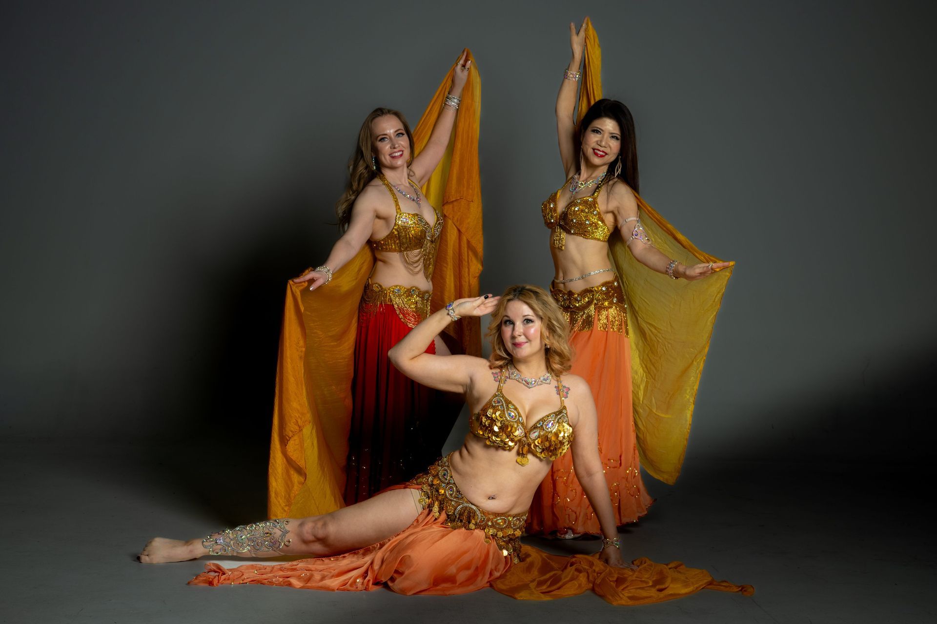 Three dancers in gold and orange costumes posing with veils against a dark studio backdrop