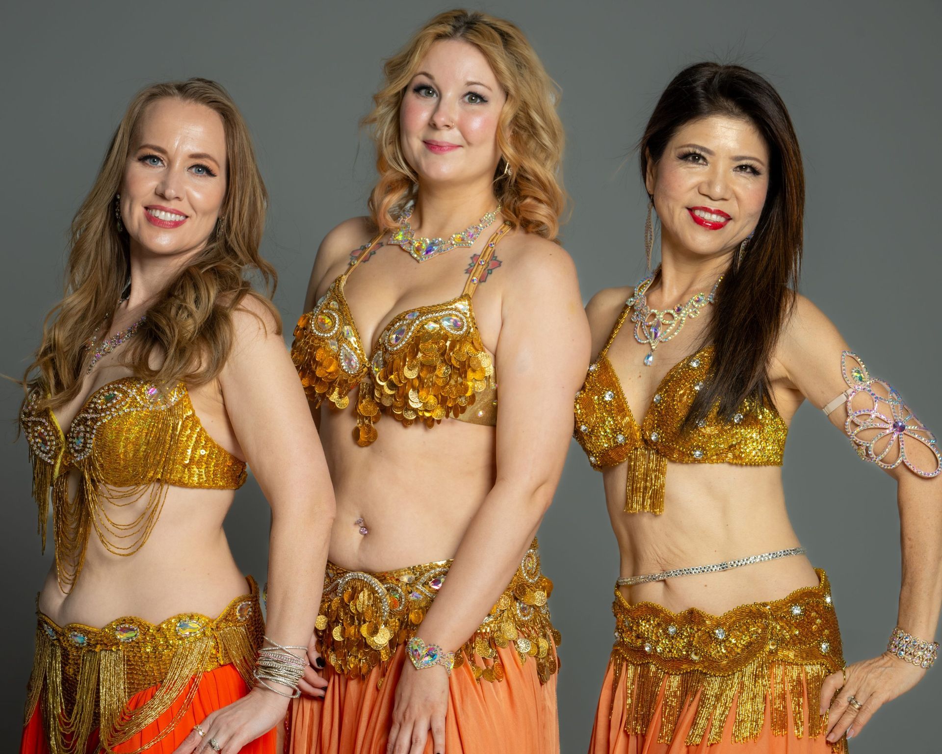 Three women in gold belly dance costumes pose against a gray backdrop.