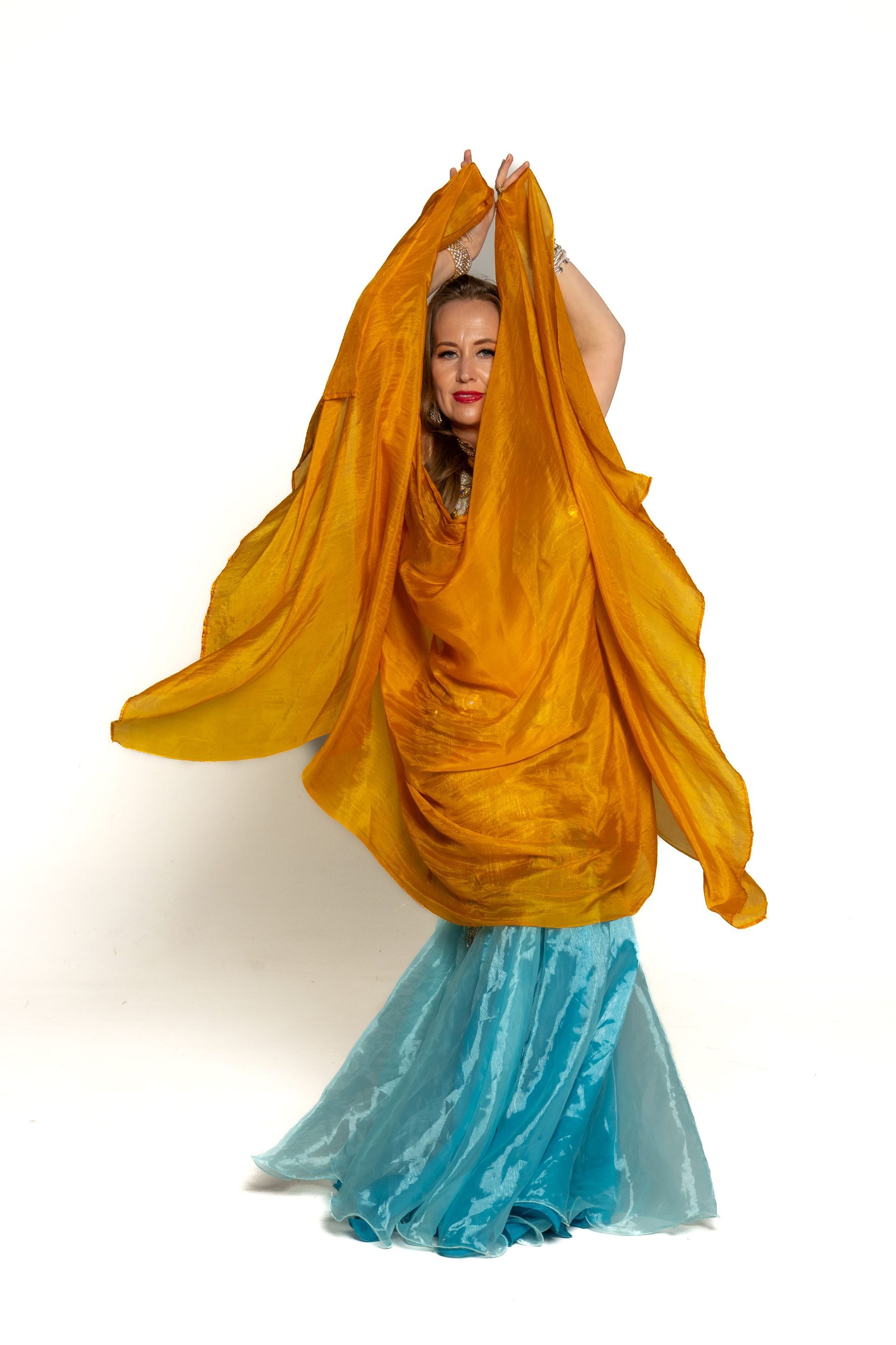 Woman in a flowing orange and turquoise outfit posing with arms raised against a white background