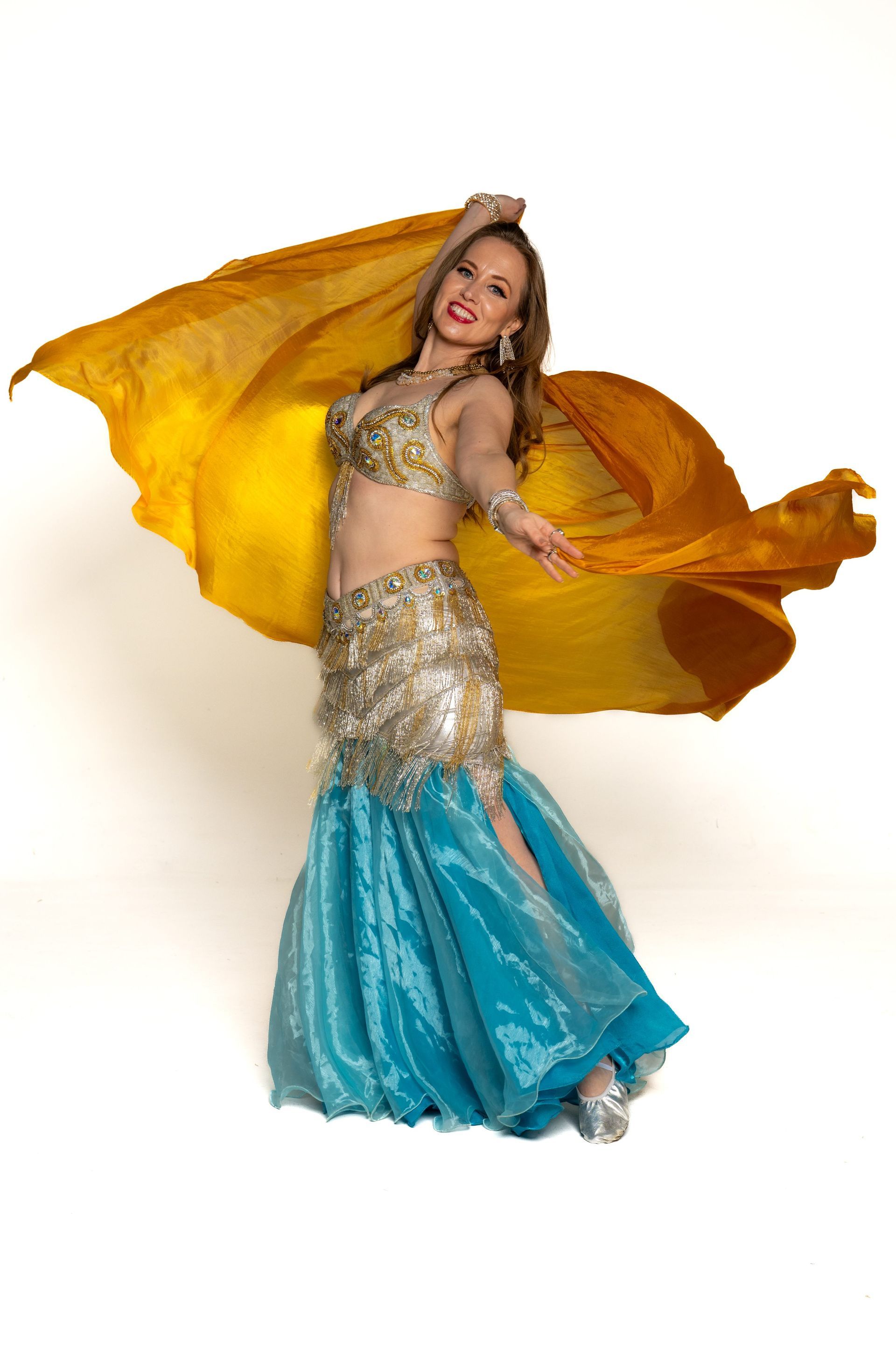 Dancer in gold and turquoise costume holding flowing yellow-orange veils against a white background