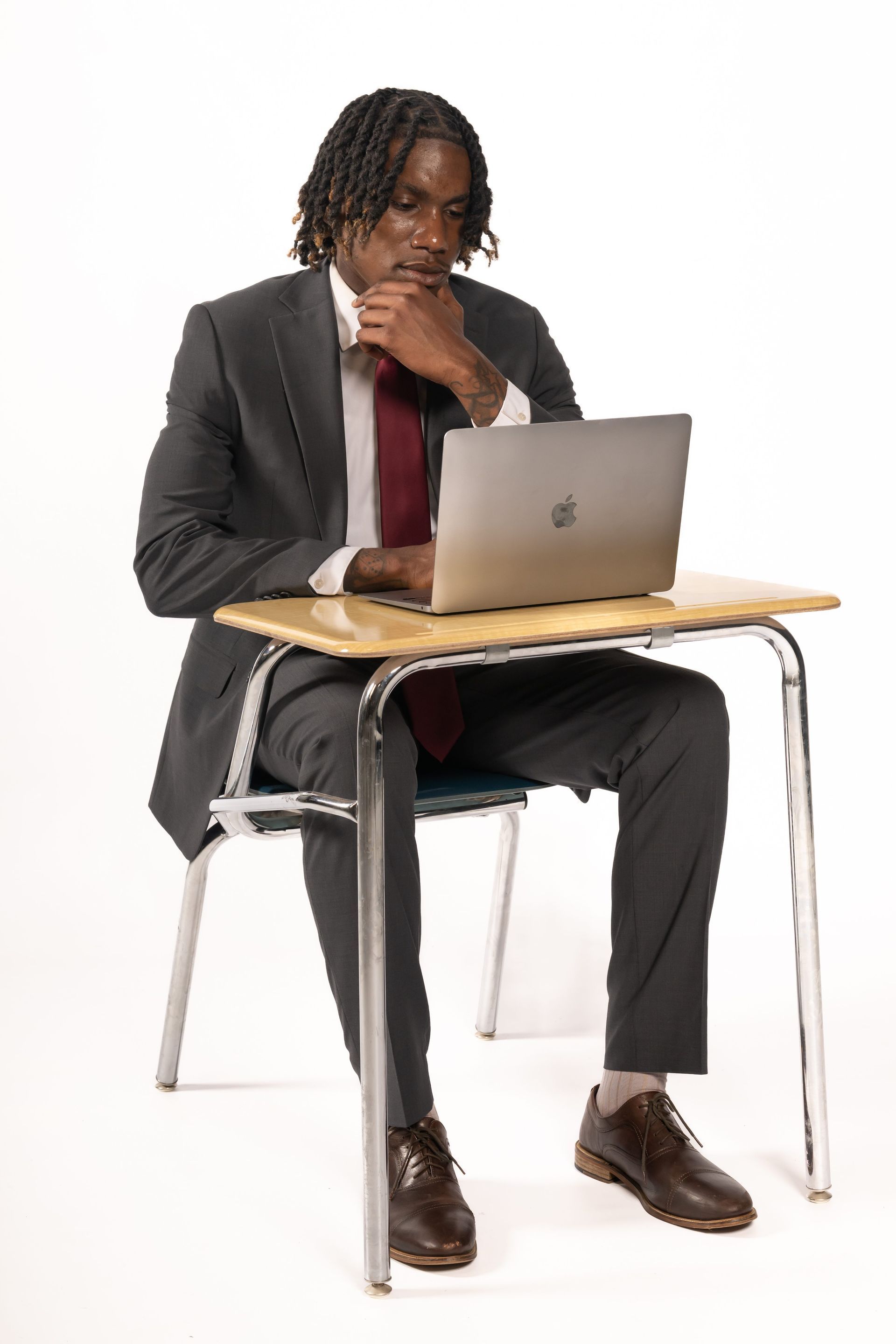 Person in a dark suit sitting at a desk with a laptop, resting their chin on their hand.