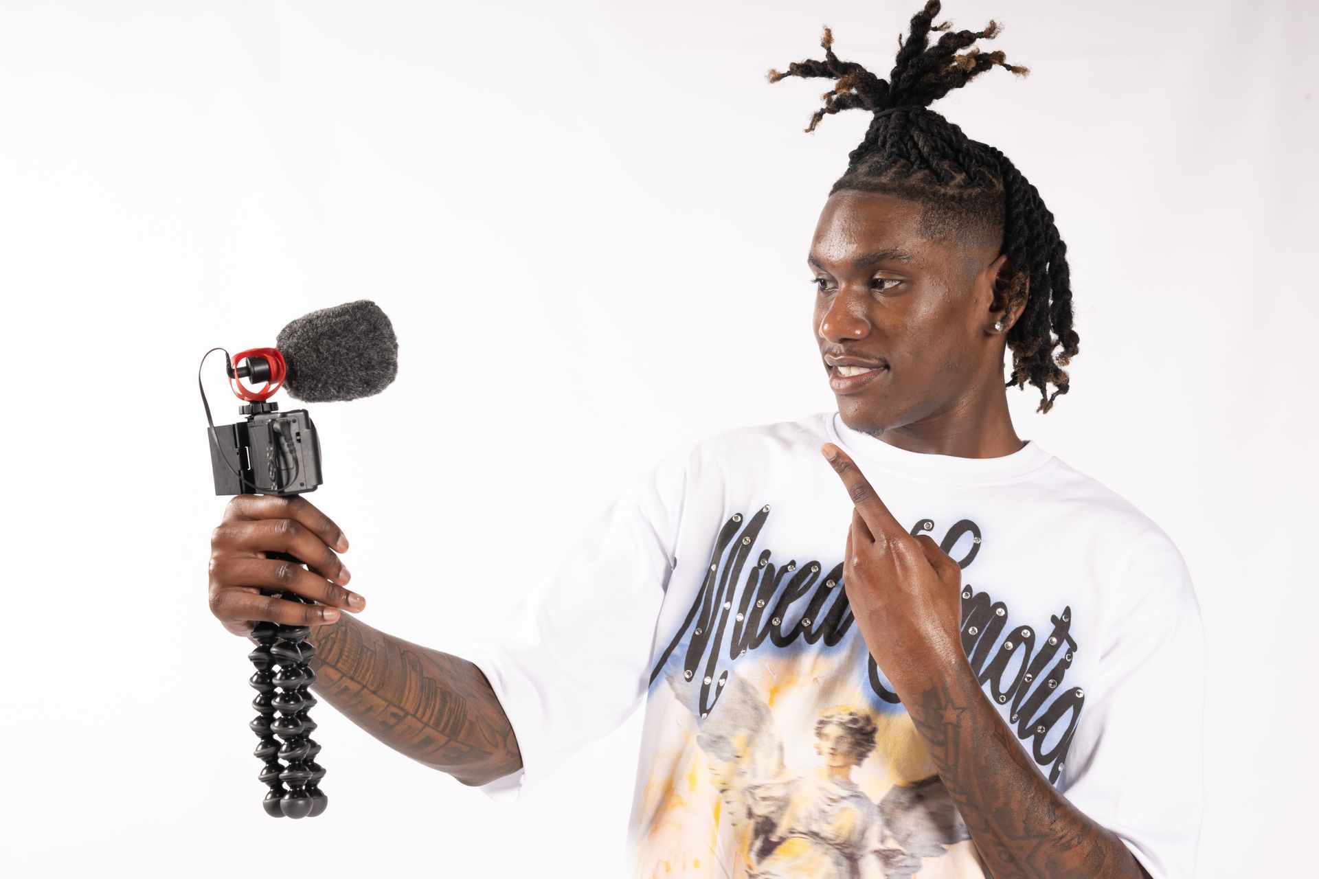 Man holding a handheld camera with microphone while speaking against a white background