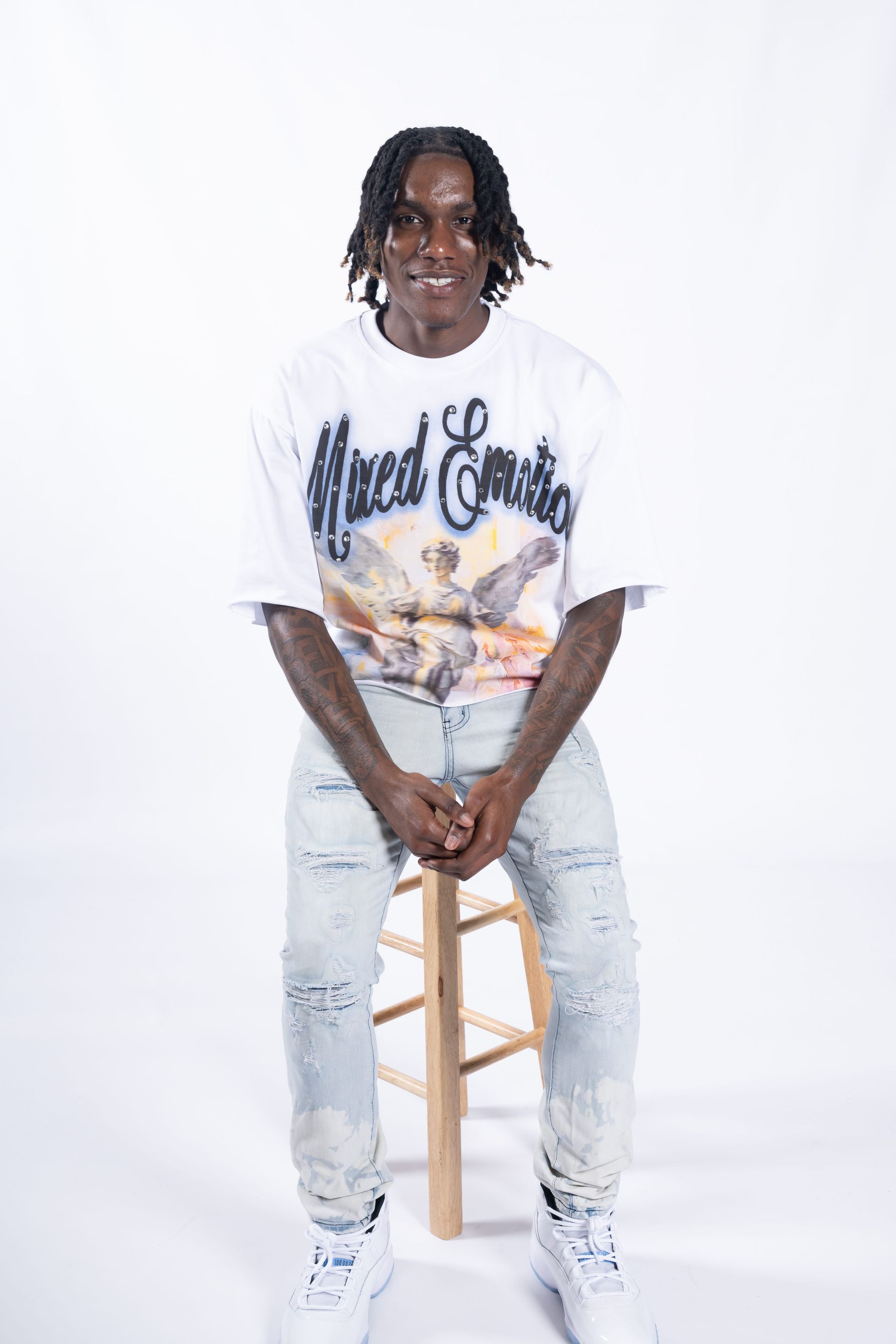 Person sitting on a stool in a white studio, wearing a graphic tee, light jeans, and white sneakers.