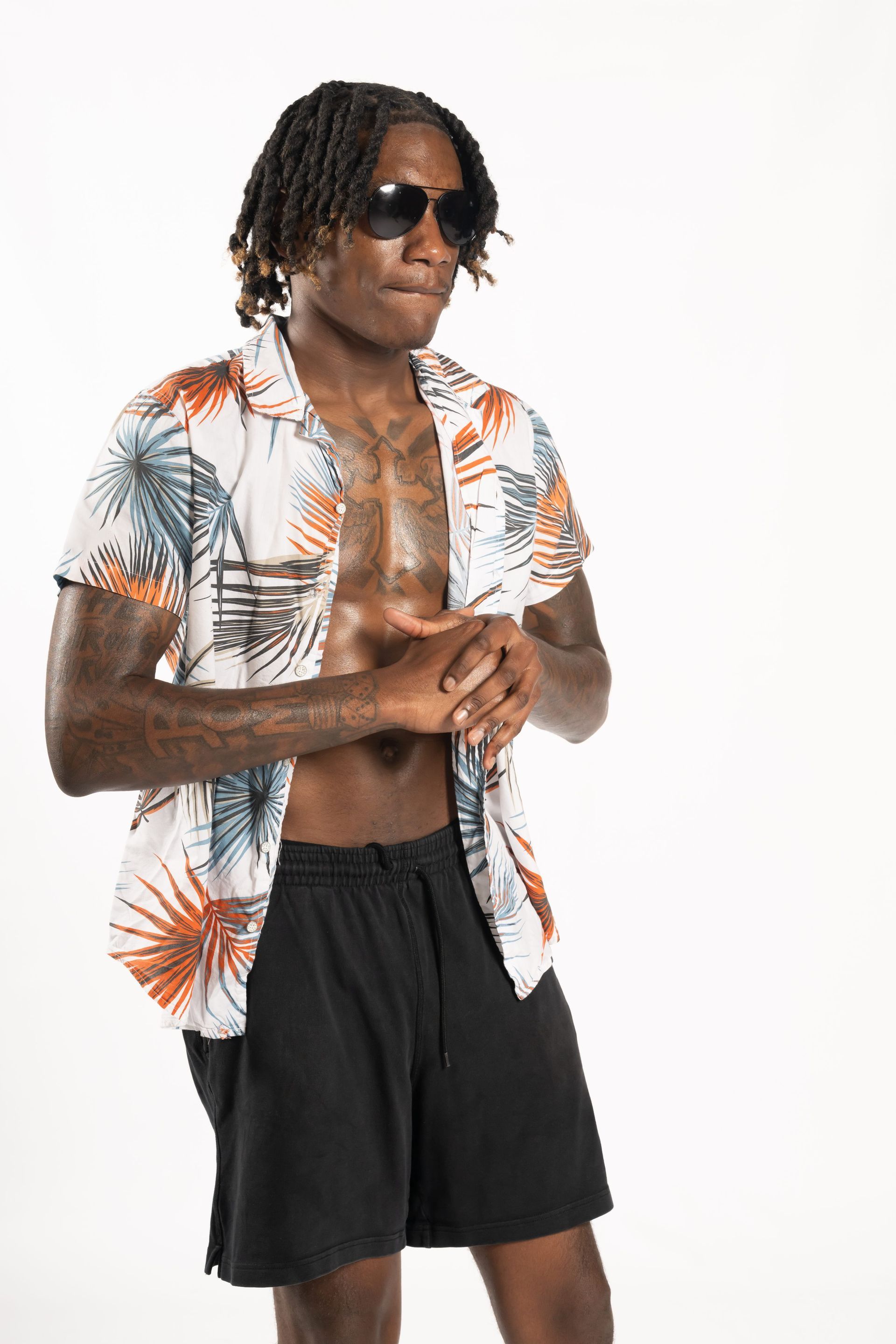 Man in sunglasses wearing an open tropical shirt and black shorts, standing against a white background.