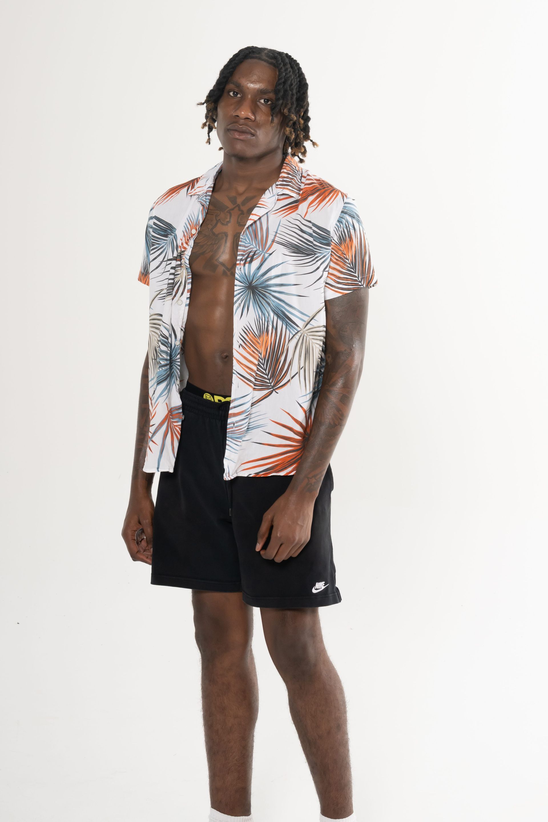 Man in an open tropical shirt, black shorts, and white sneakers standing against a white background