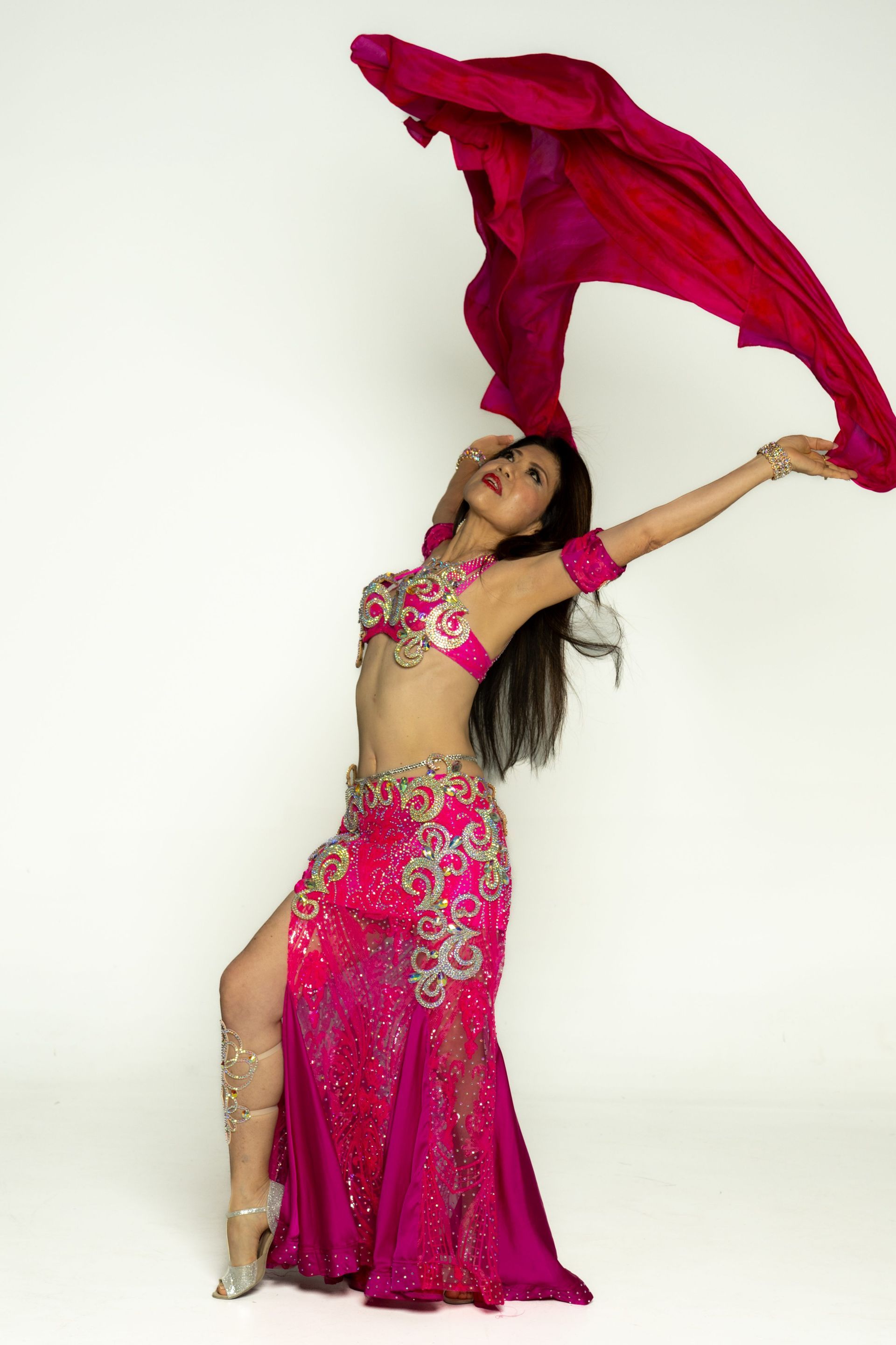 Belly dancer in pink costume holding a flowing red veil overhead, posed against a plain white background