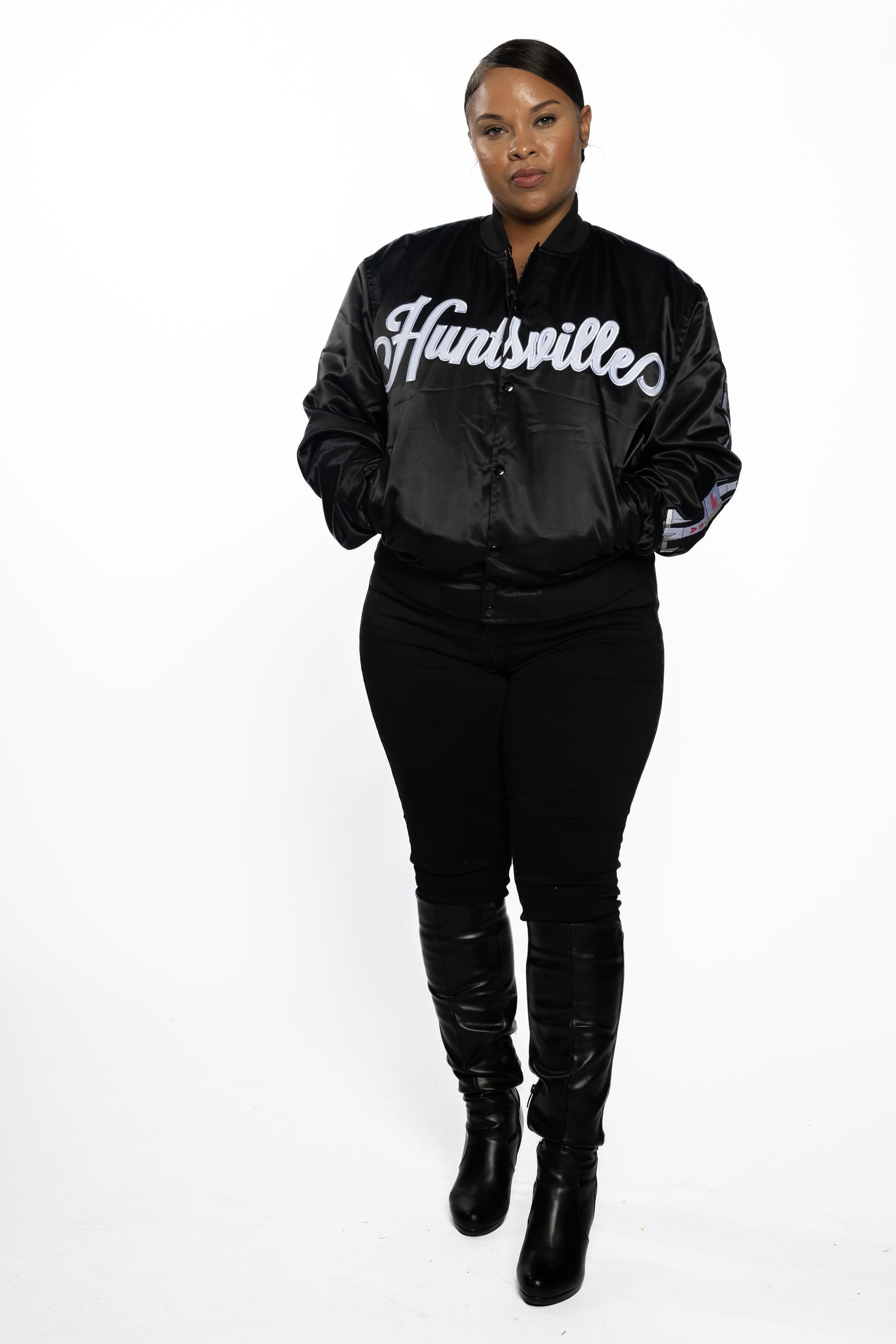 Person in a black jacket and pants standing against a white background, hands in pockets.