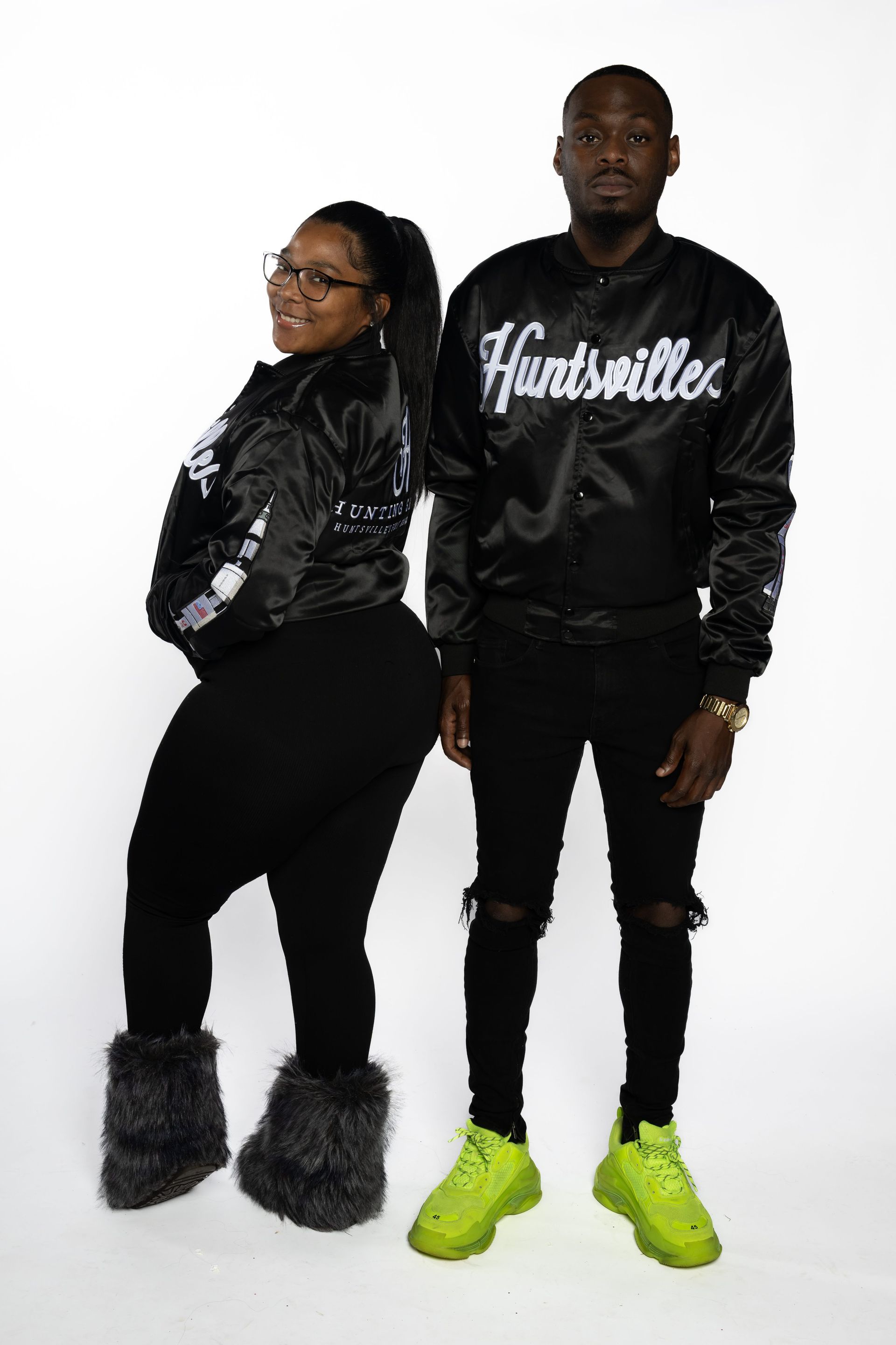 Two people pose in black outfits; one wears bright green sneakers and the other furry boots.